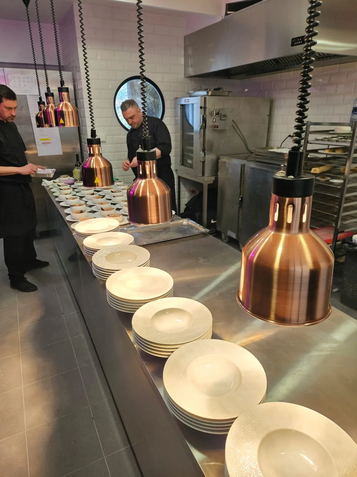 Deux chefs en cuisine préparant des plats devant des assiettes alignées sous des lampes en cuivre suspendues.