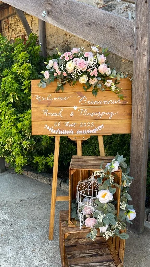 Un panneau en bois avec des fleurs roses et blanches au sommet, affichant un message de bienvenue en anglais et en français, avec des détails pour un événement daté du 06 août 2022, placé devant des arbustes verts et une structure en bois.