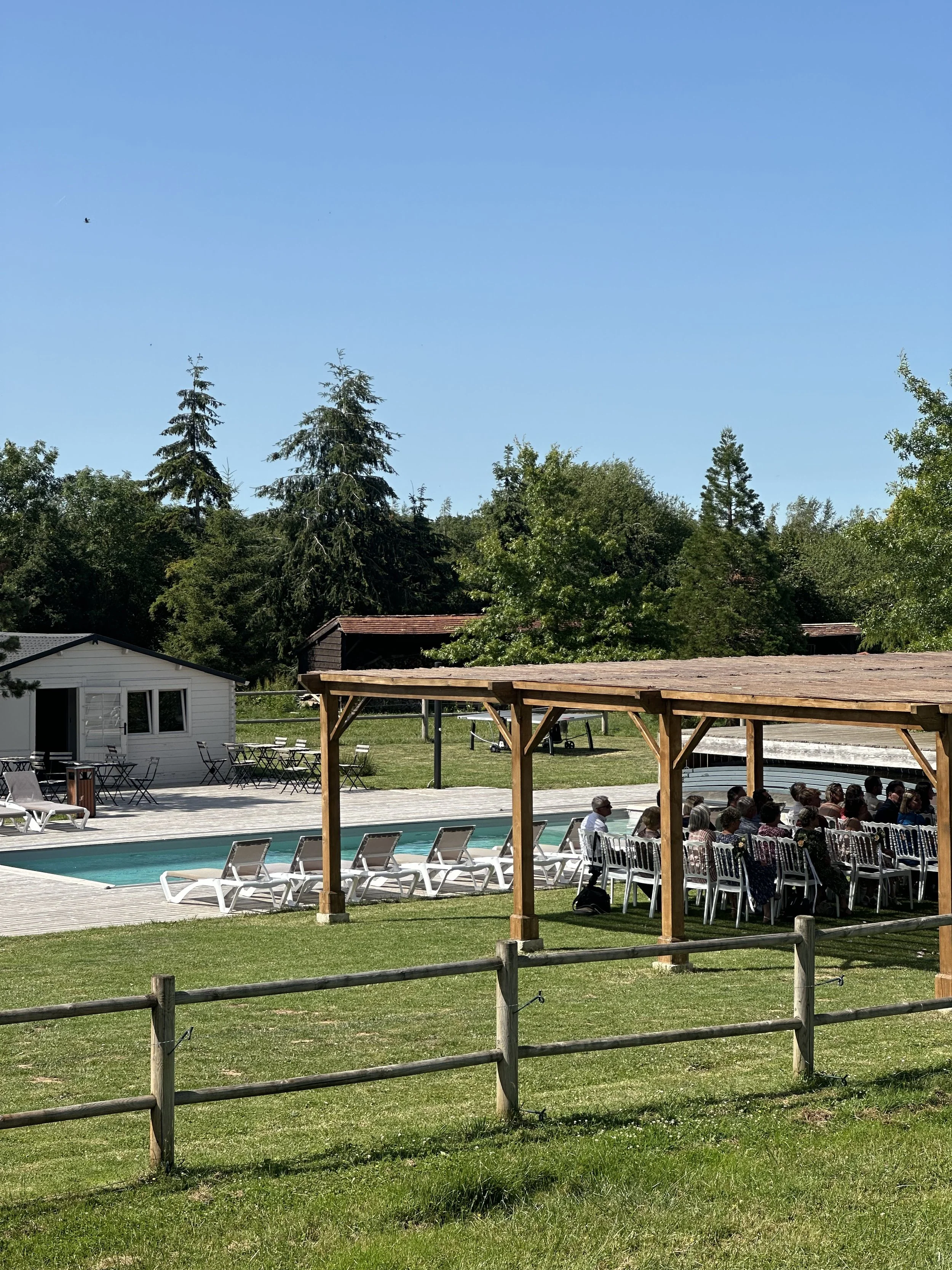 Une piscine extérieure avec des transats blancs autour, un espace ombragé avec des chaises où des personnes sont assises, un bâtiment blanc, une clôture en bois, et un ciel clair avec quelques arbres verts en arrière-plan.