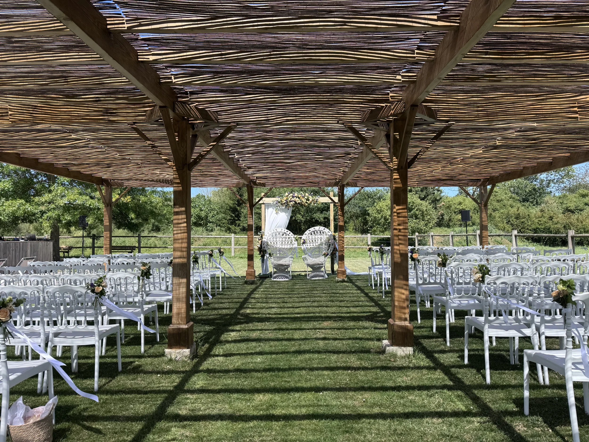 Une décoration de mariage en extérieur avec un gazebo en bois, des chaises blanches, des fleurs et une toile de fond verte avec des arbres et un ciel bleu.
