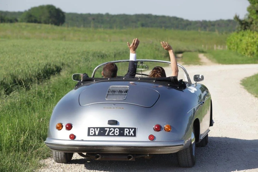 Une voiture décapotable grise sur une route de campagne, avec deux personnes à l'intérieur, levant les bras en l'air, sous un ciel clair et verdoyant.