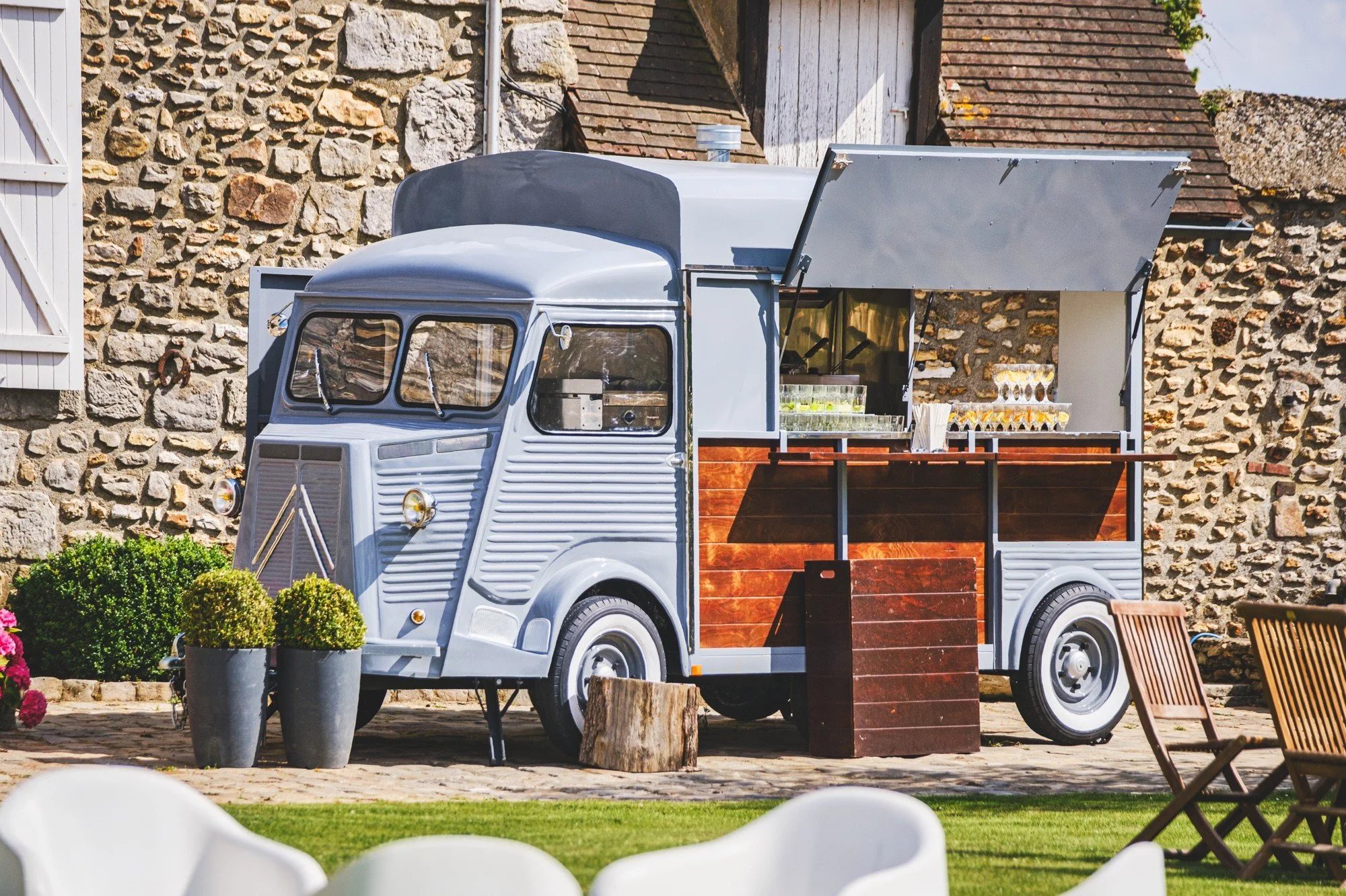 Un camion de restauration de style vintage converti en food truck, stationné dans un espace extérieur avec des plantes en pot et des chaises en bois, devant un mur en pierre.