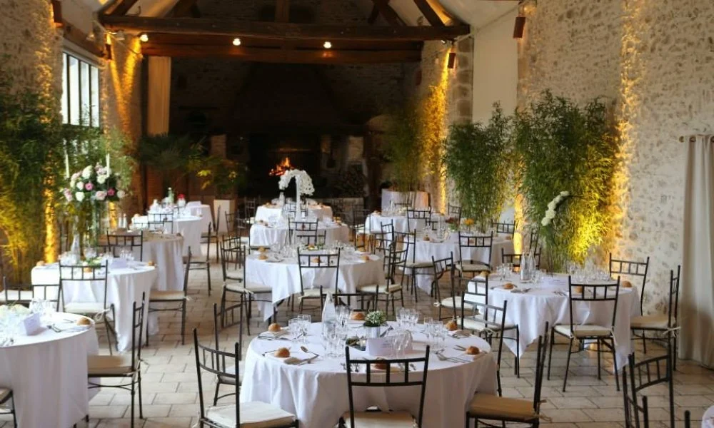 Salle de réception élégante avec tables rondes habillées de nappes blanches, décorée de bouquets de fleurs et éclairée chaleureusement, en préparation pour un événement ou un dîner.