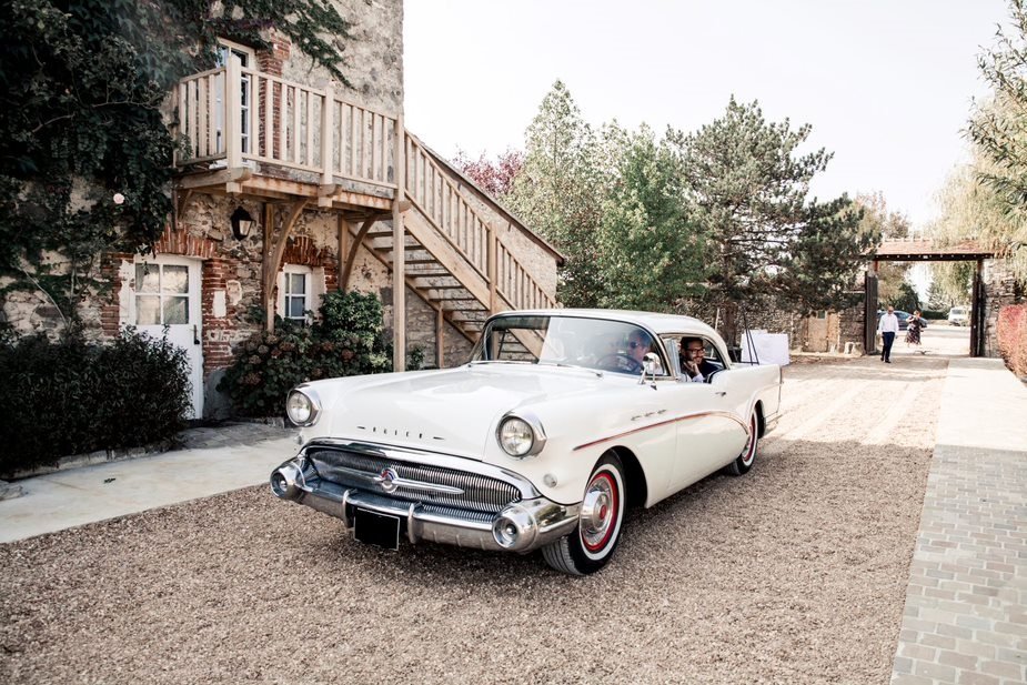 Voiture ancienne blanche garée dans une cour, avec deux personnes à l'intérieur. En arrière-plan, un bâtiment en pierre avec une escalier en bois, et des arbres et un portail en arrière-plan.