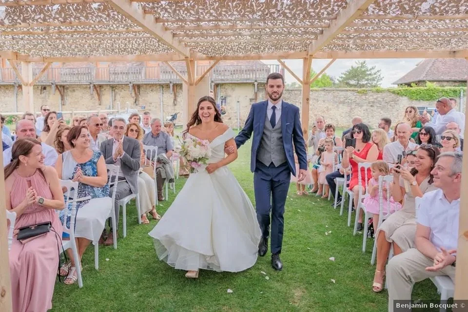 Un couple de mariés marche dans une allée lors d'une cérémonie en plein air, entouré d'invités assis et applaudissant, sous une pergola en bois décorée.