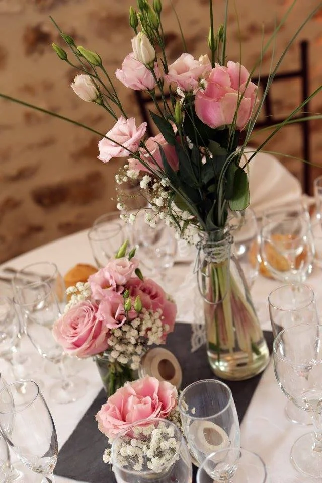 Un arrangement floral avec des roses roses, des épis de lisianthus blancs, des petits œillets blancs, et des branches d'eucalyptus dans une bouteille en verre sur une table de fête. Autour, des verres à vin et un décor de fête.