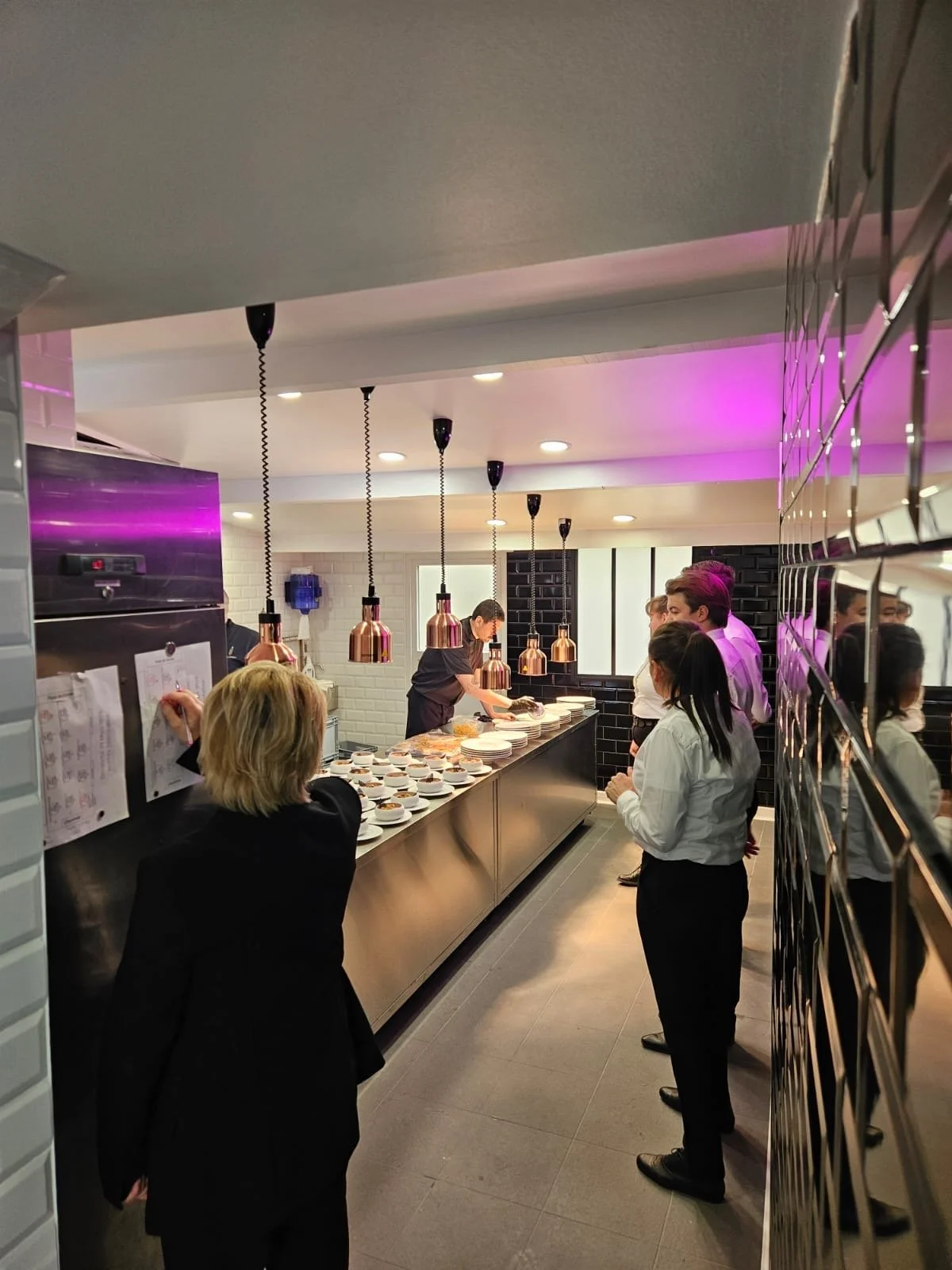 Une scène dans un restaurant ou une salle de dégustation, où plusieurs personnes attendent leur repas ou leur dégustation, avec un chef ou un serveur préparant des plats derrière le comptoir.