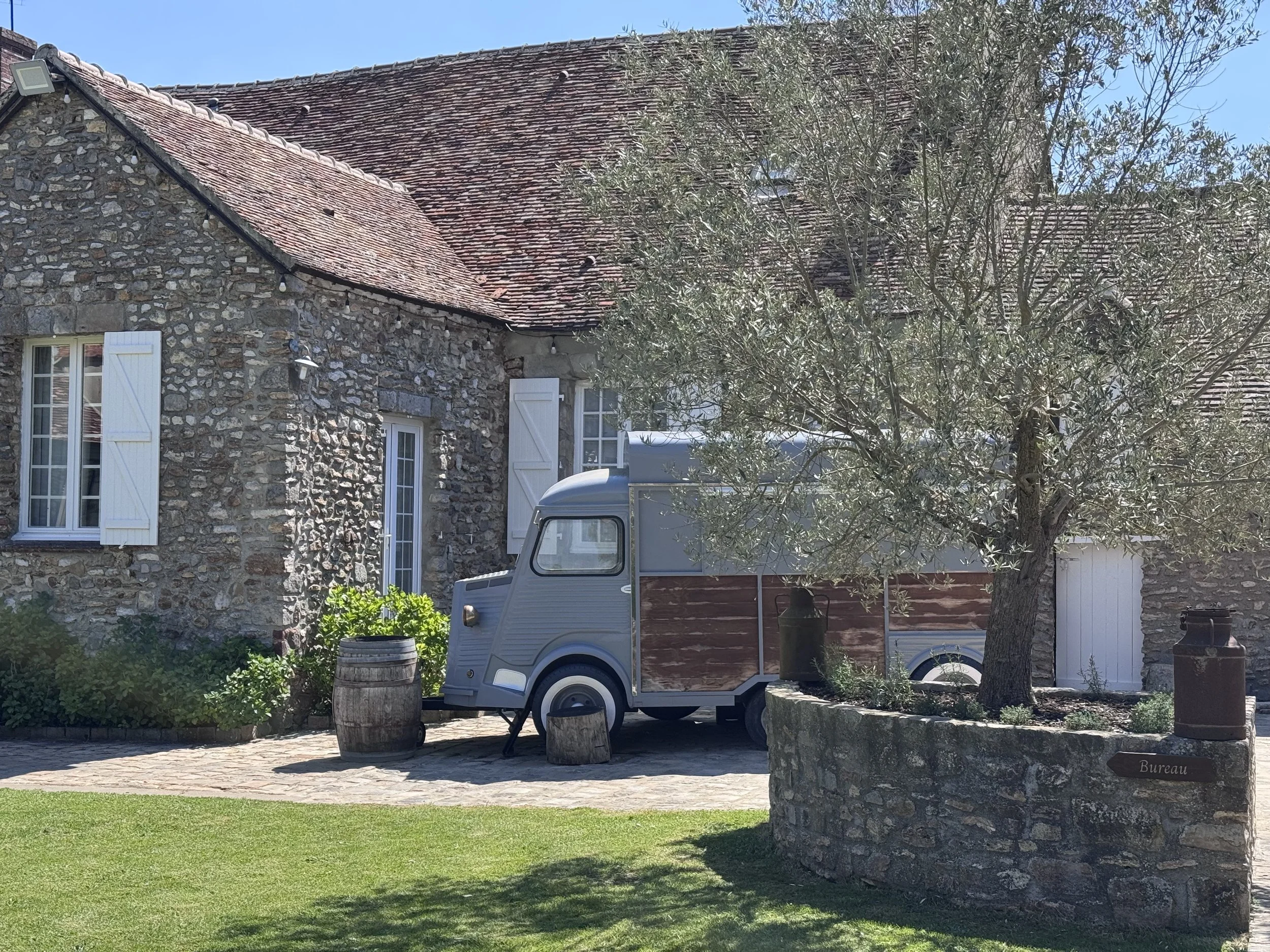 Maison en pierre avec toit en tuiles rouges, fenêtres à volets blancs, arbre dans le jardin, petite voiture rétro gris, baril en bois, un panier de légumes, et un panneau indiquant 'Bureau' dans un jardin ensoleillé avec herbe verte.