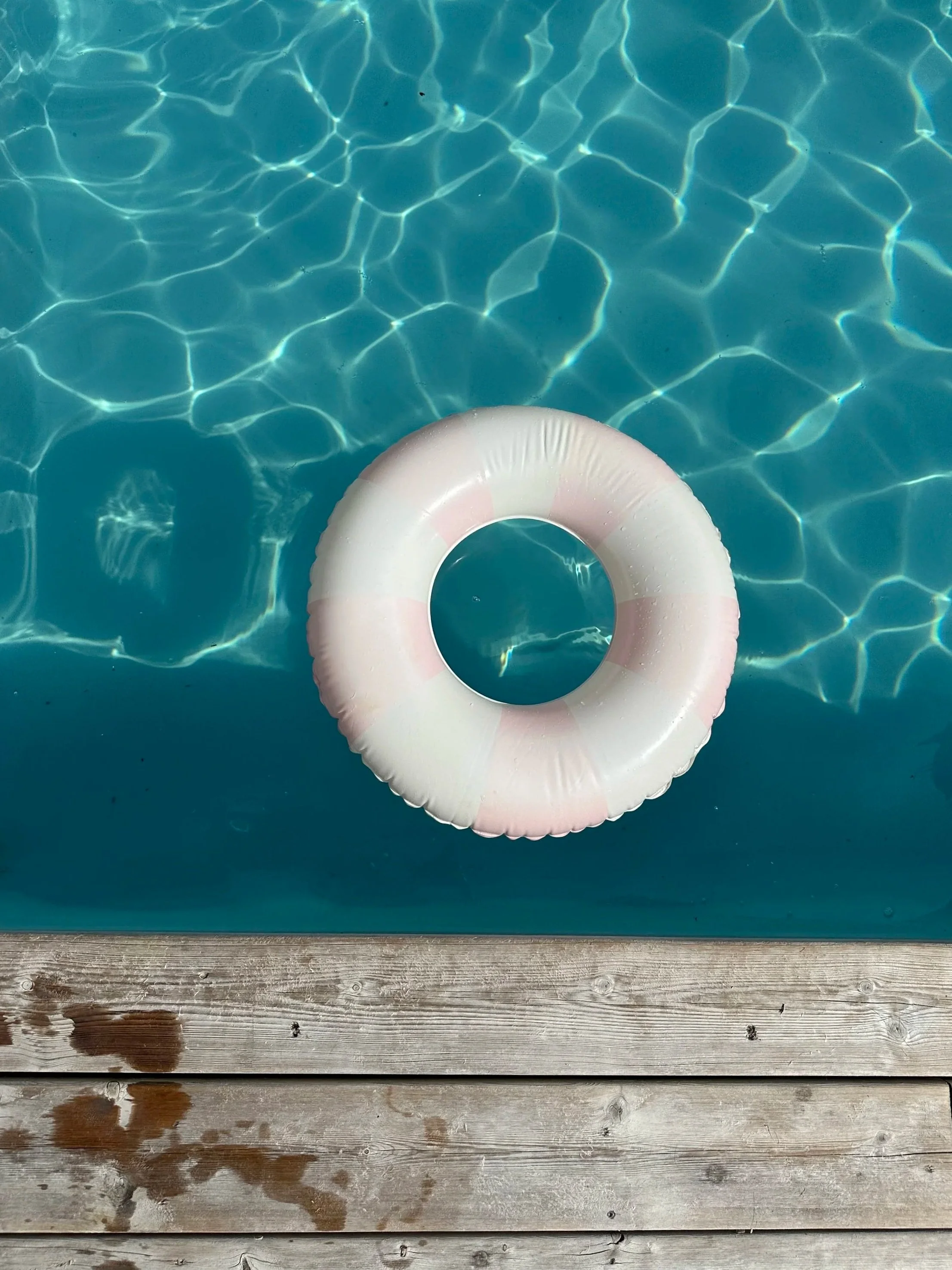 Une bouée en forme de cercle flotte sur une piscine d'eau bleue avec des reflets lumineux. La partie inférieure de l'image montre un pont en bois avec des marques d'eau et des taches de pluie.