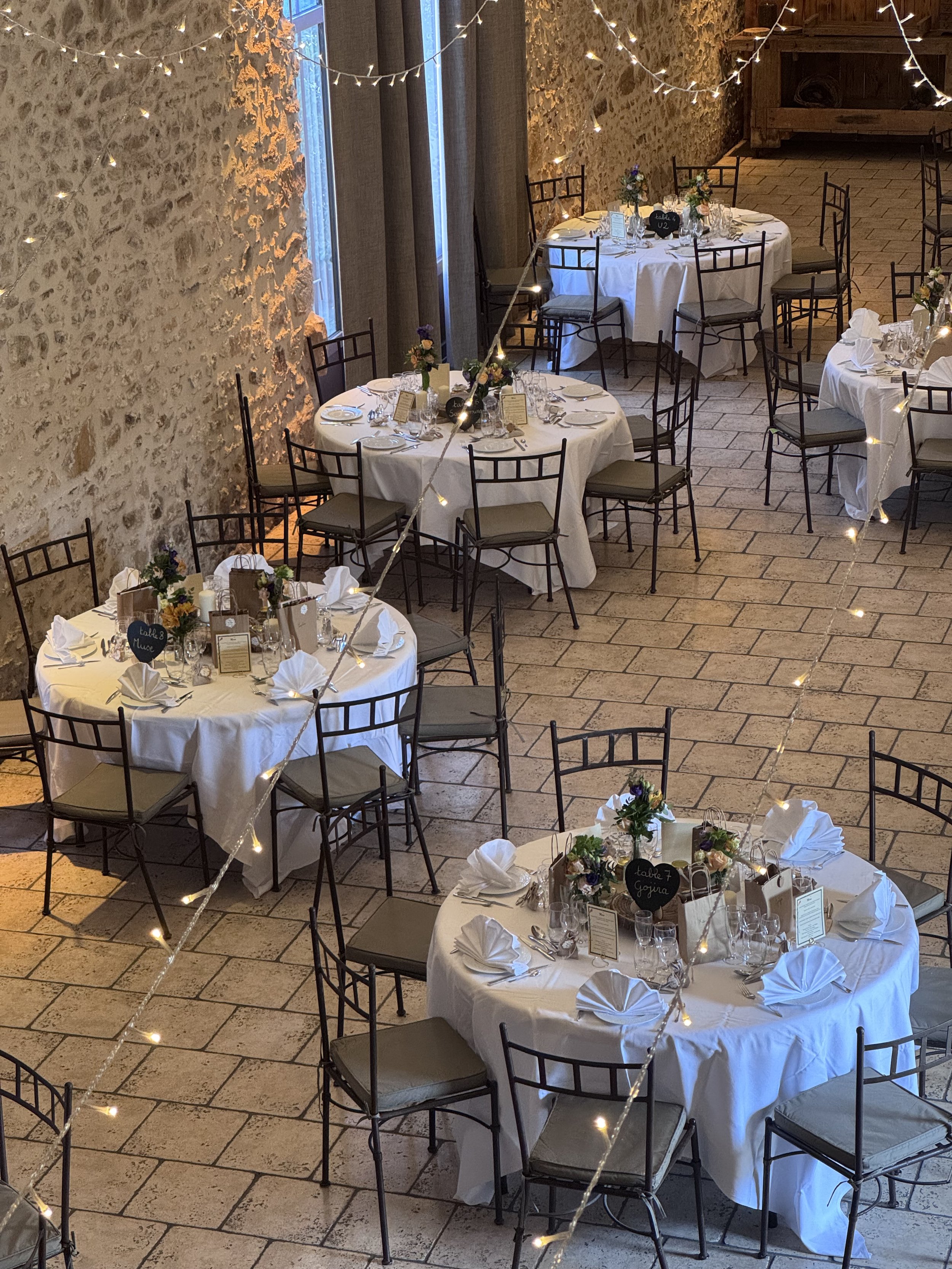 Salle de réception décorée pour un événement, avec tables rondes vêtues de nappes blanches, vaisselle, fleurs, et guirlandes lumineuses suspendues, murs en pierre et éclairage chaleureux.