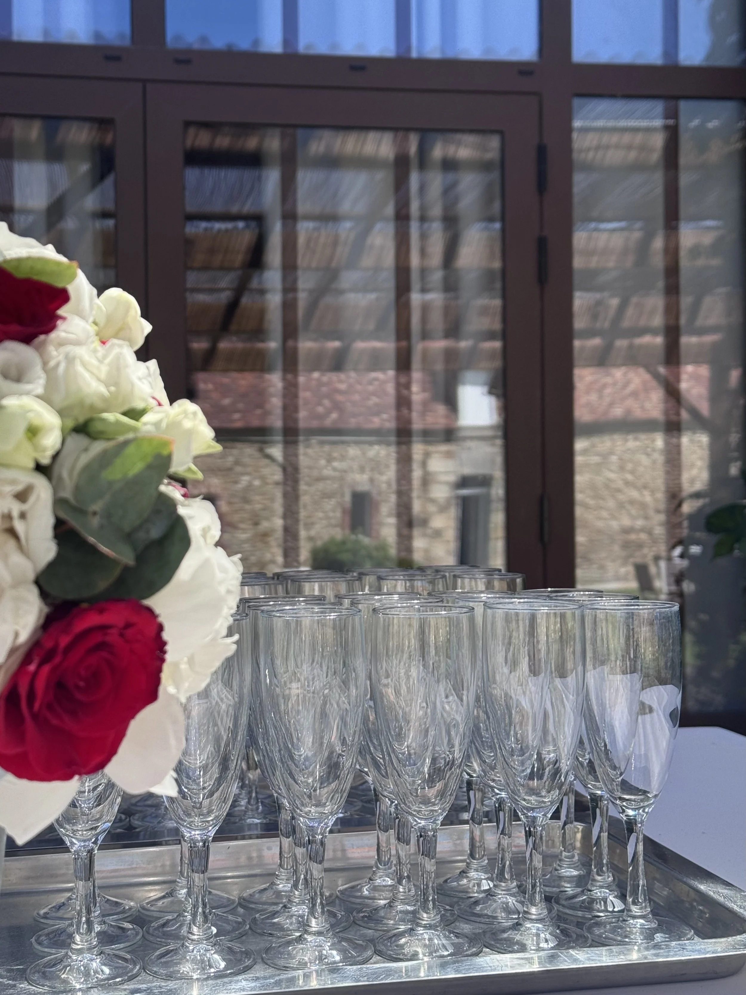 Verres en cristal vides disposés sur un plateau en métal, bouquet de fleurs blanches et rouges en premier plan et fenêtre en arrière-plan