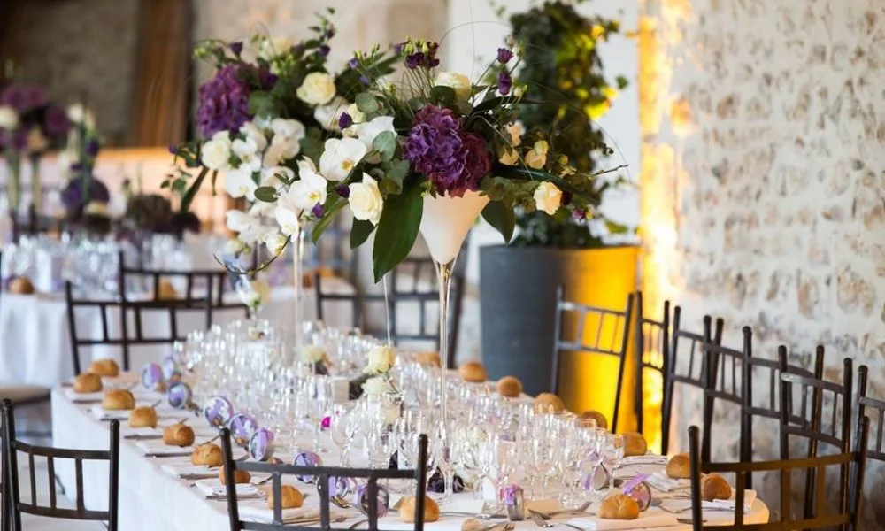Table de fête décorée avec une grande composition florale centrale et des assiettes avec des petits pains, tables et chaises pour un événement formel.