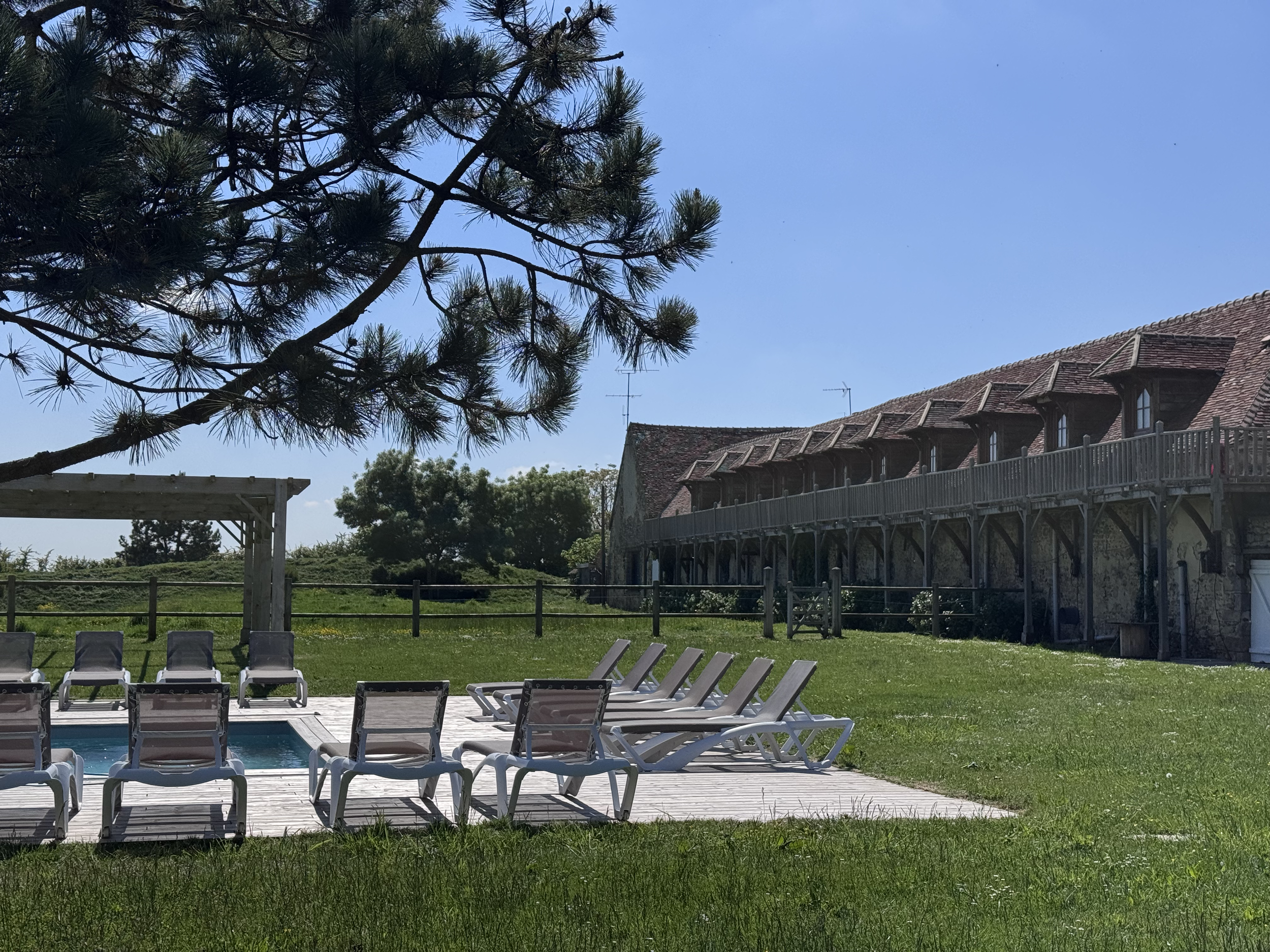 Une piscine entourée de chaises longues en plastique blanc et d'une pelouse verte, avec une maison en pierre à toiture en tuiles rouges en arrière-plan, sous un ciel bleu clair.