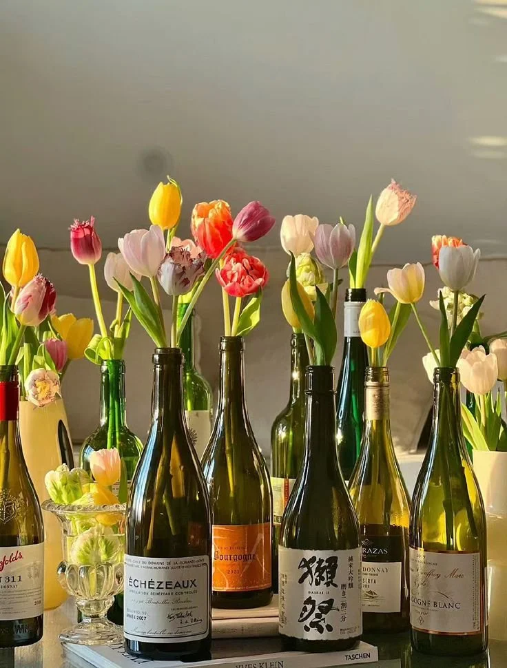 Vases de bouteilles en verre contenant des tulipes roses, jaunes, rouges, blanches et violettes sur une surface, avec un fond neutre.
