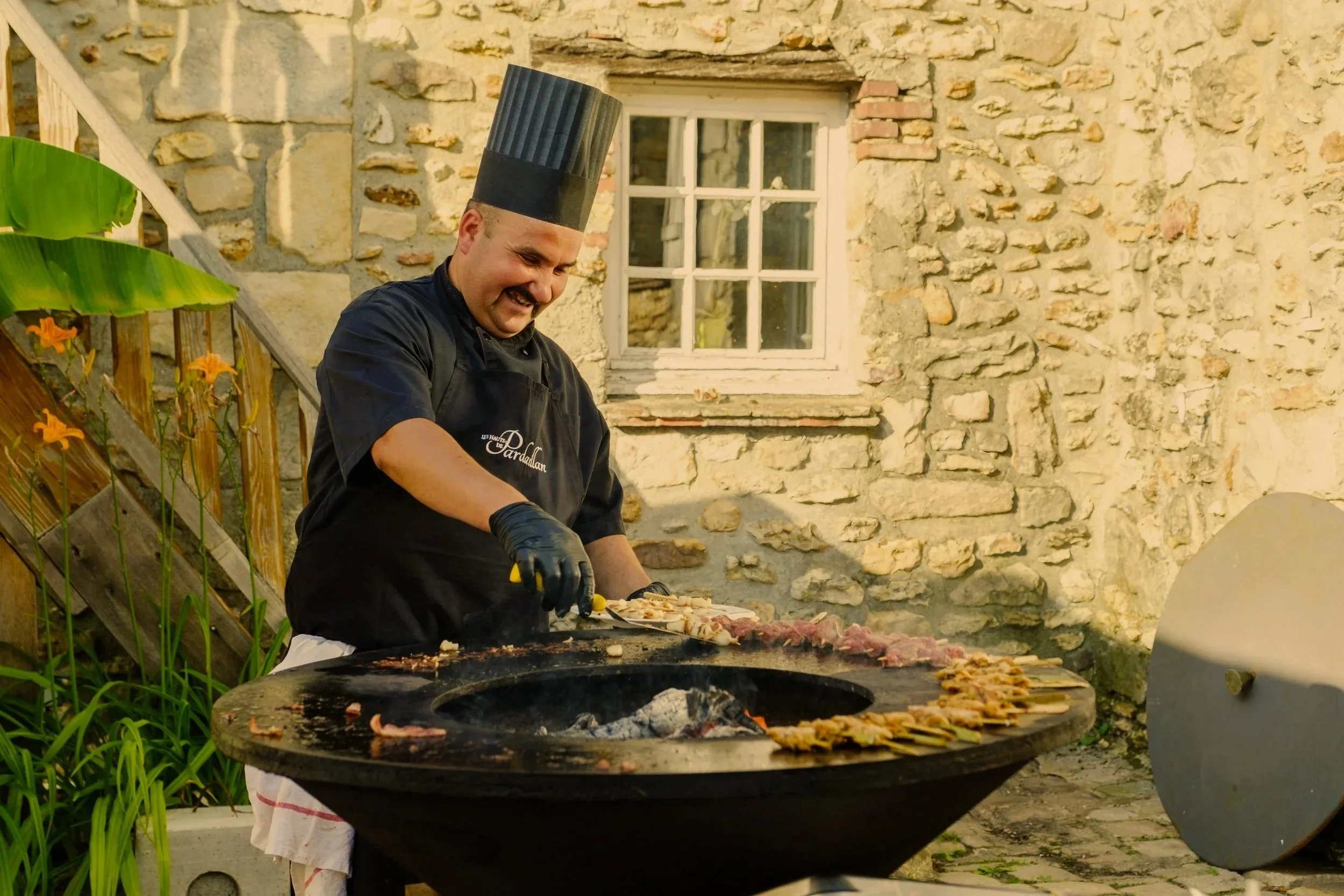 Un chef en train de préparer des pizzas sur une grande surface de cuisson extérieure en pierre, avec un mur de pierre en arrière-plan.