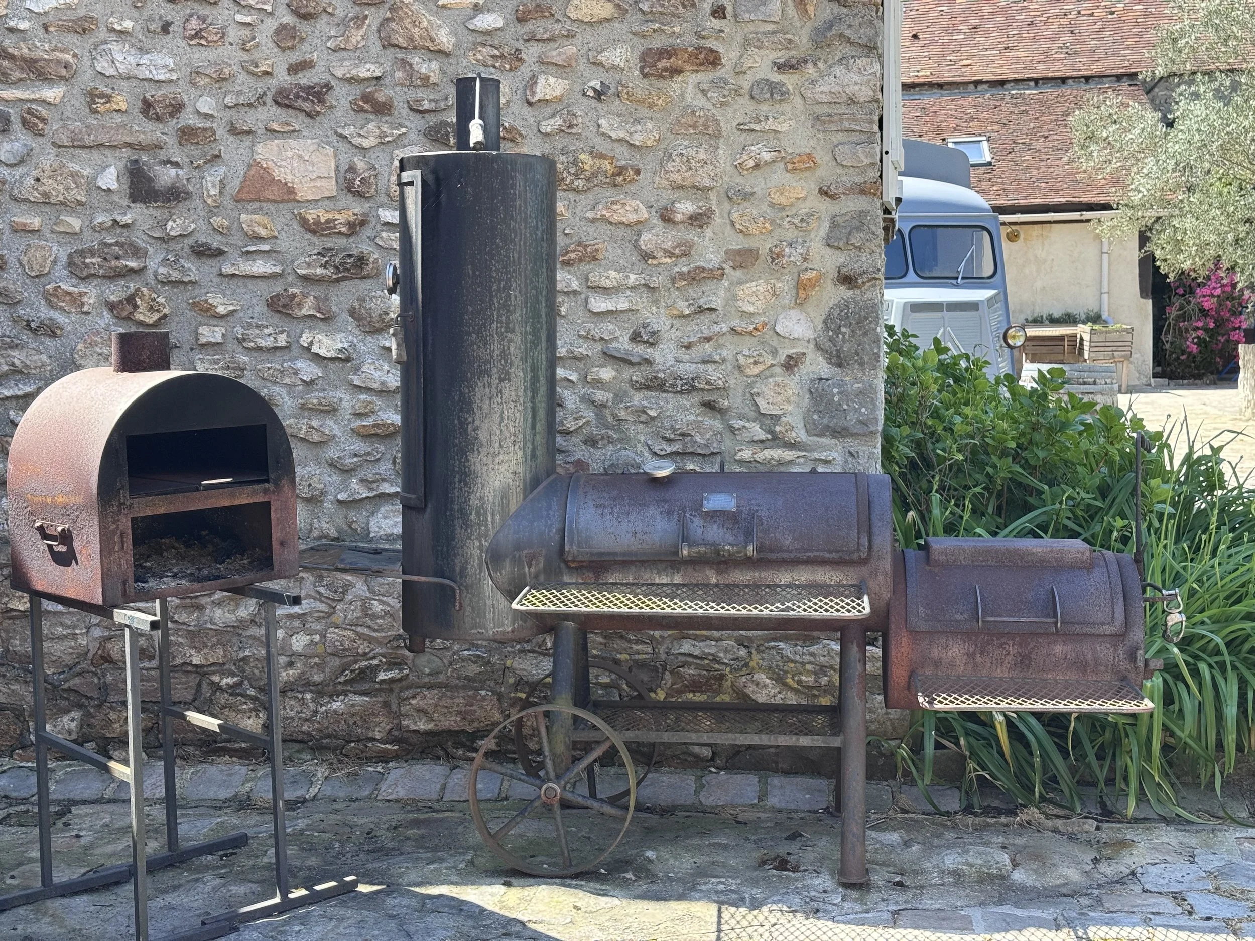 Ancien barbecue en métal rouillé, face à un mur en pierre, avec une maison en arrière-plan et un arbuste à droite.