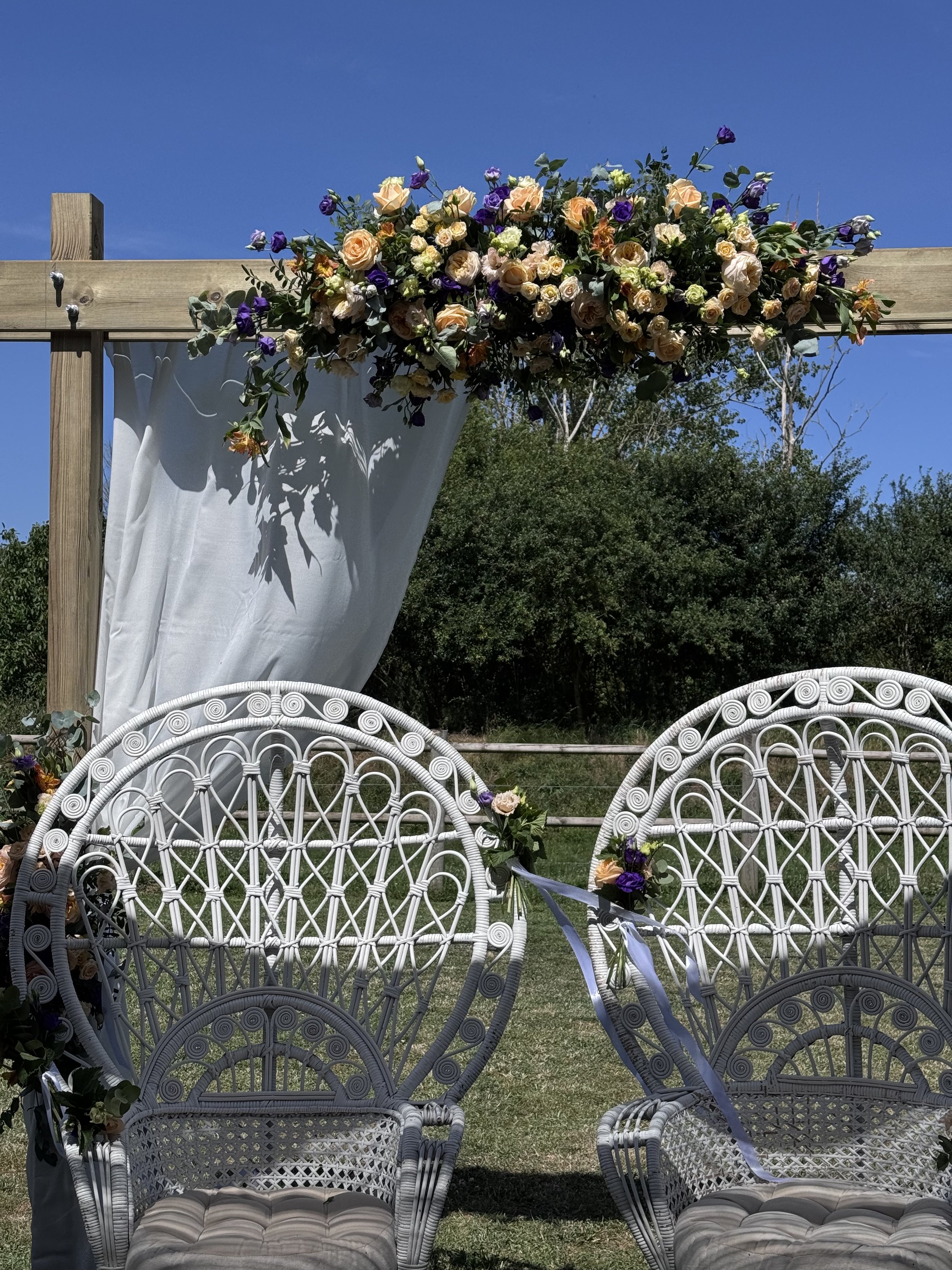 Une arche décorée de fleurs, avec des chaises blanches en osier au premier plan, dans un cadre extérieur avec un ciel bleu clair.