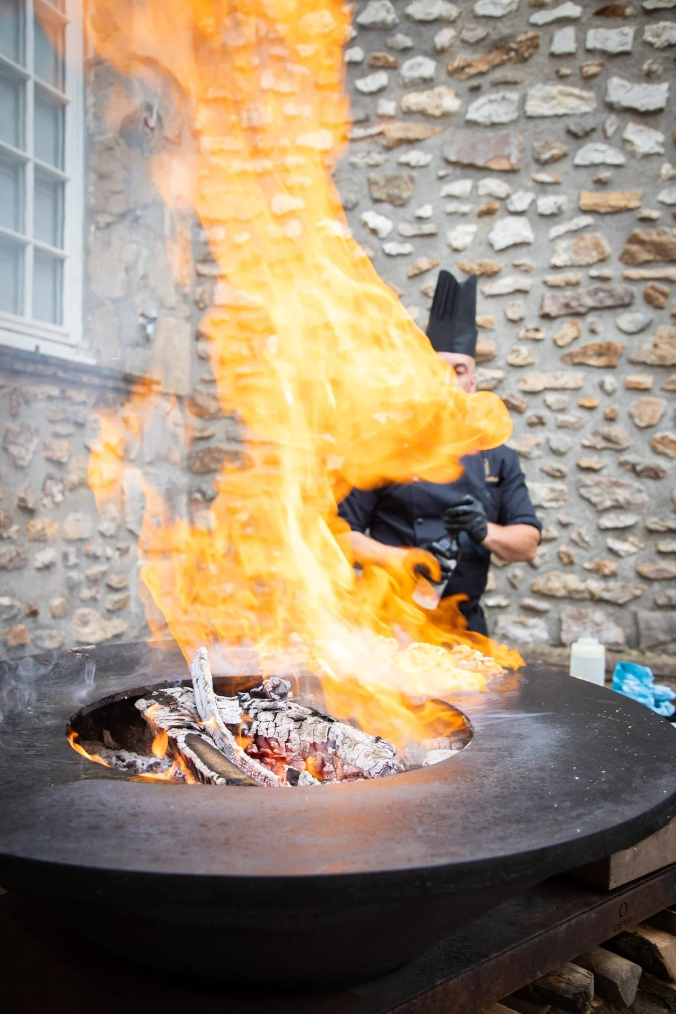 Un chef ou cuisinier en noir, portant un chapeau de chef, à proximité d'une flamme d'un feu de bois ou de barbecue, avec un mur de pierre en arrière-plan.