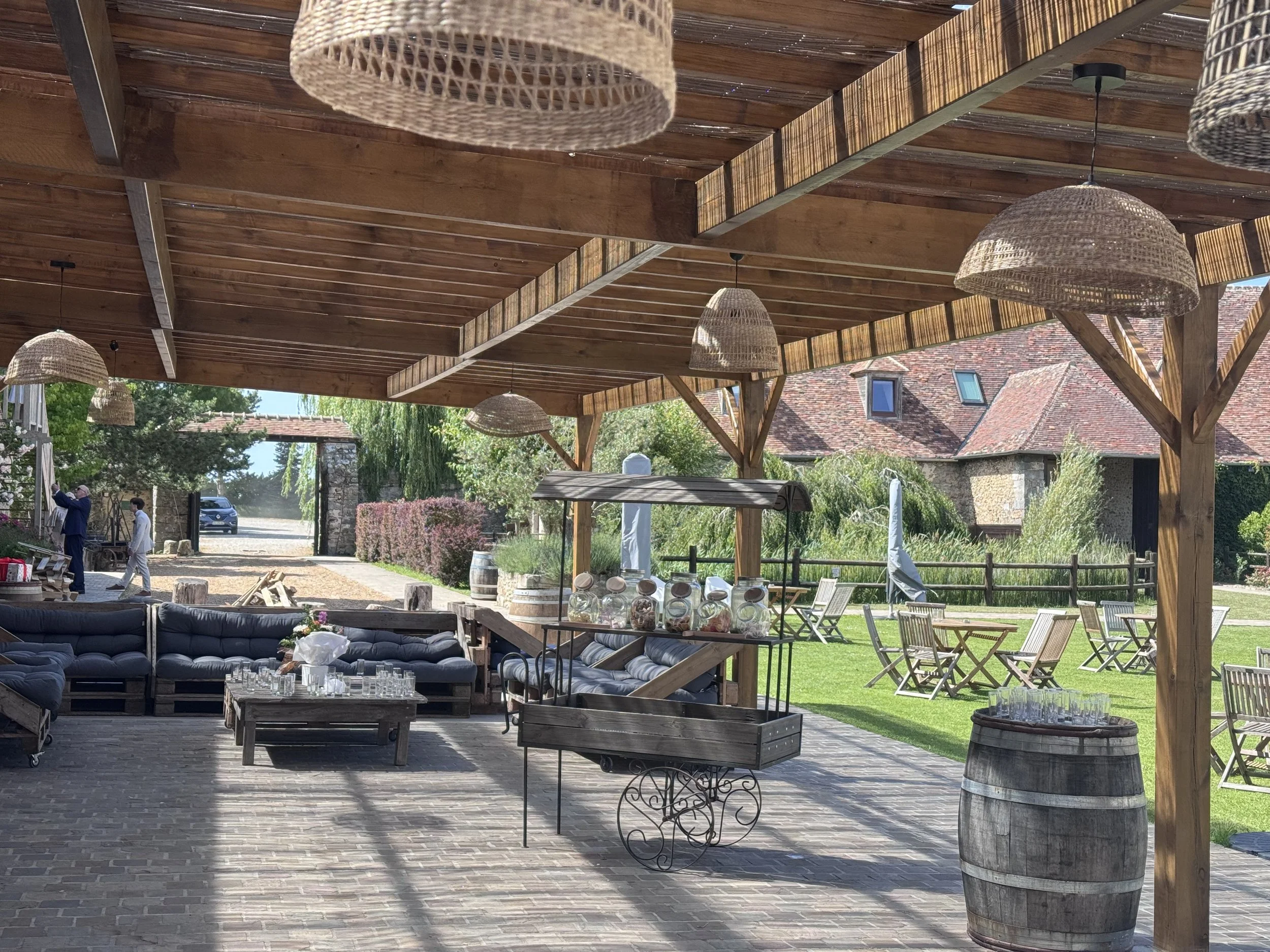 Une terrasse couverte en bois avec des lampes suspensions en osier, des meubles en rotin avec des coussins, et des chaises pliantes en bois, dans un cadre extérieur avec des bâtiments en pierre et un jardin verdoyant.