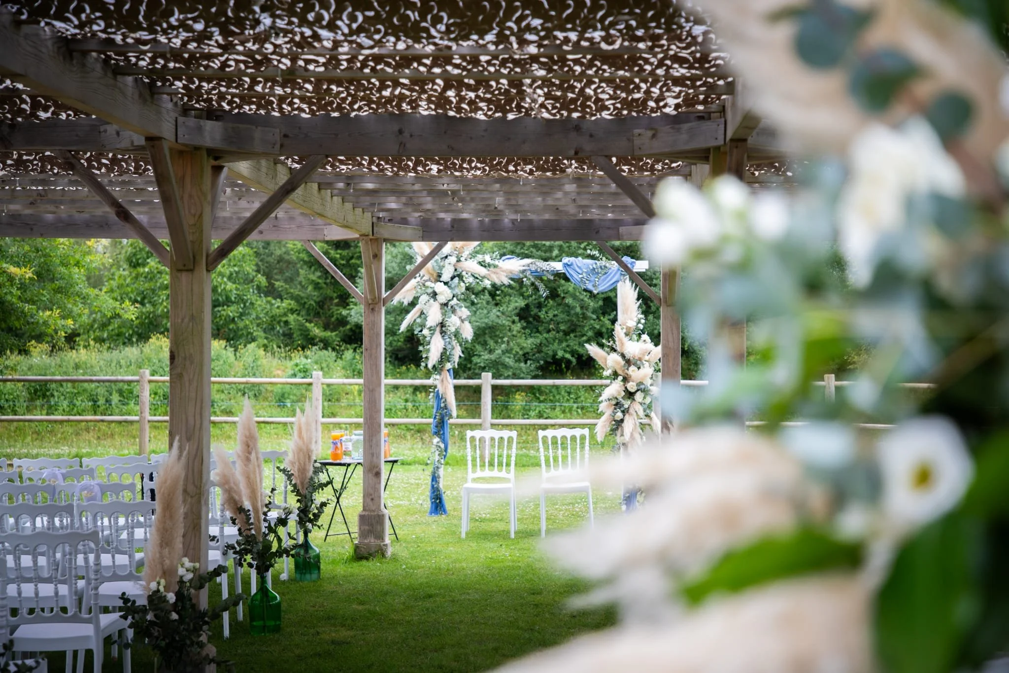Une scène d'extérieur aménagée pour une célébration, avec des chaises blanches, des décorations florales et un plafond en bois, dans un environnement vert avec des arbres.