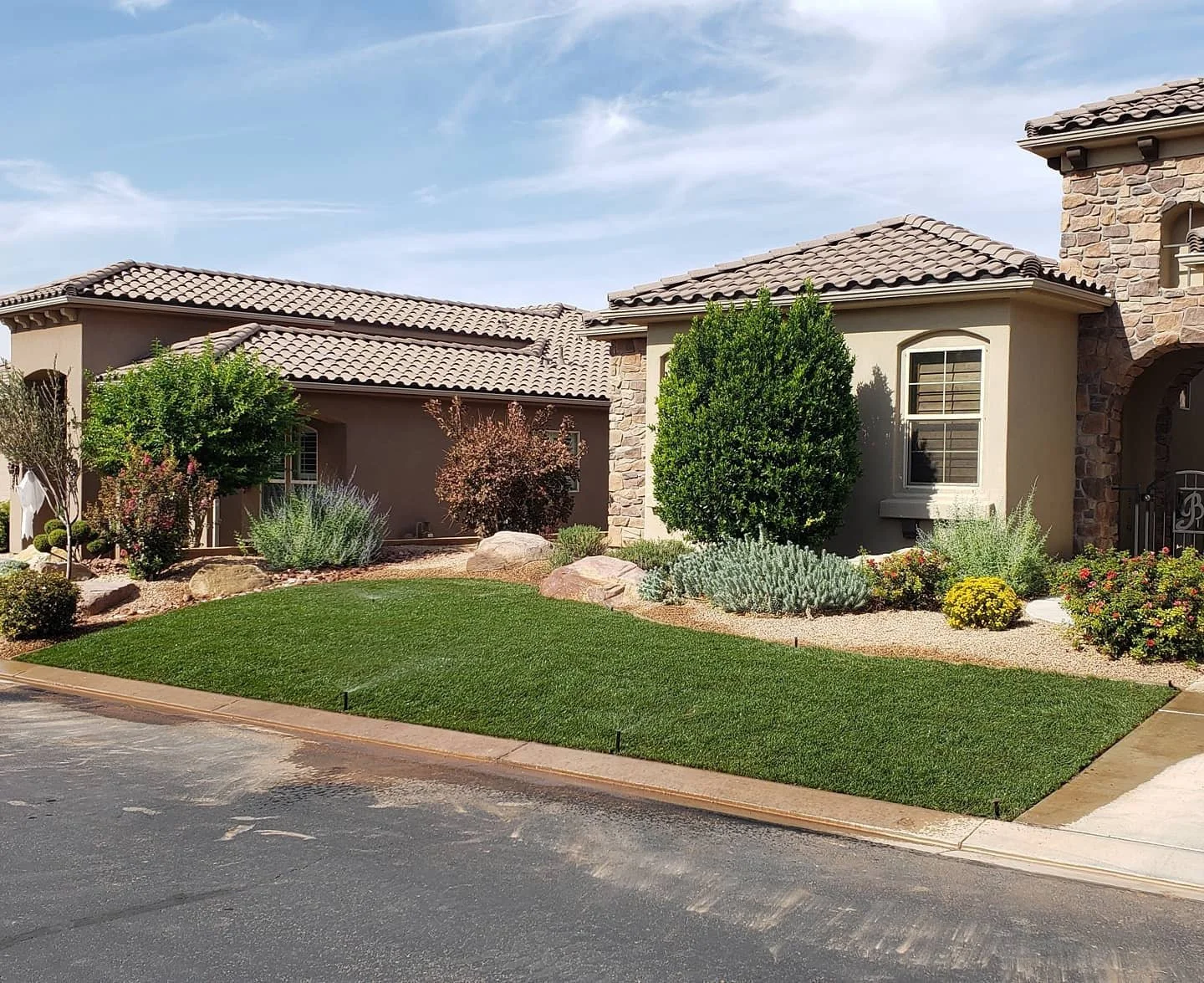 A residential house with a well-maintained front yard featuring green grass, bushes, and small trees under a partly cloudy sky.