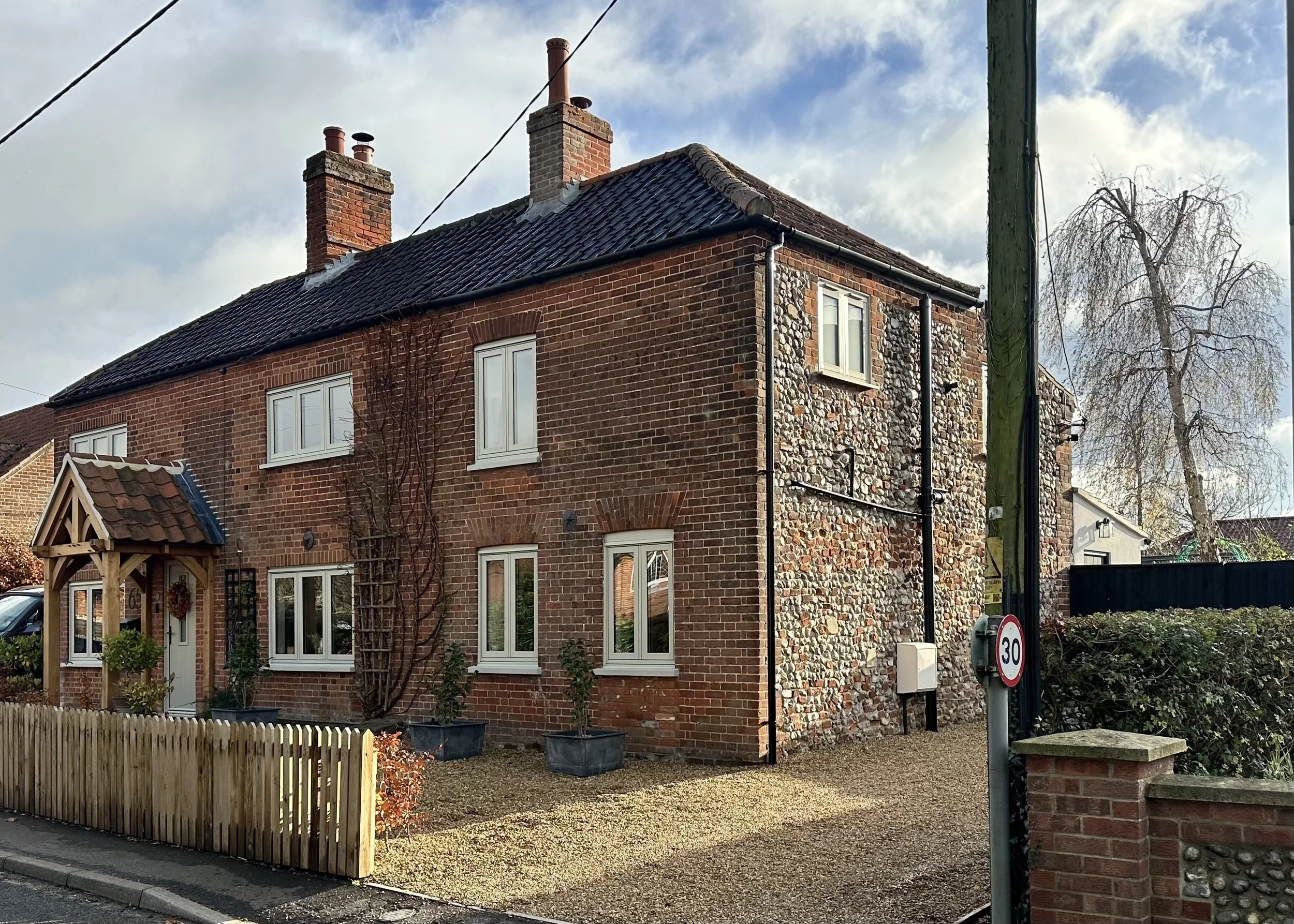 A two-story brick house with a tiled roof, multiple white-framed windows, a small porch with a tiled roof, a wooden fence, and a gravel yard with potted plants. There is a speed limit sign of 30 mph and a utility pole on the street.