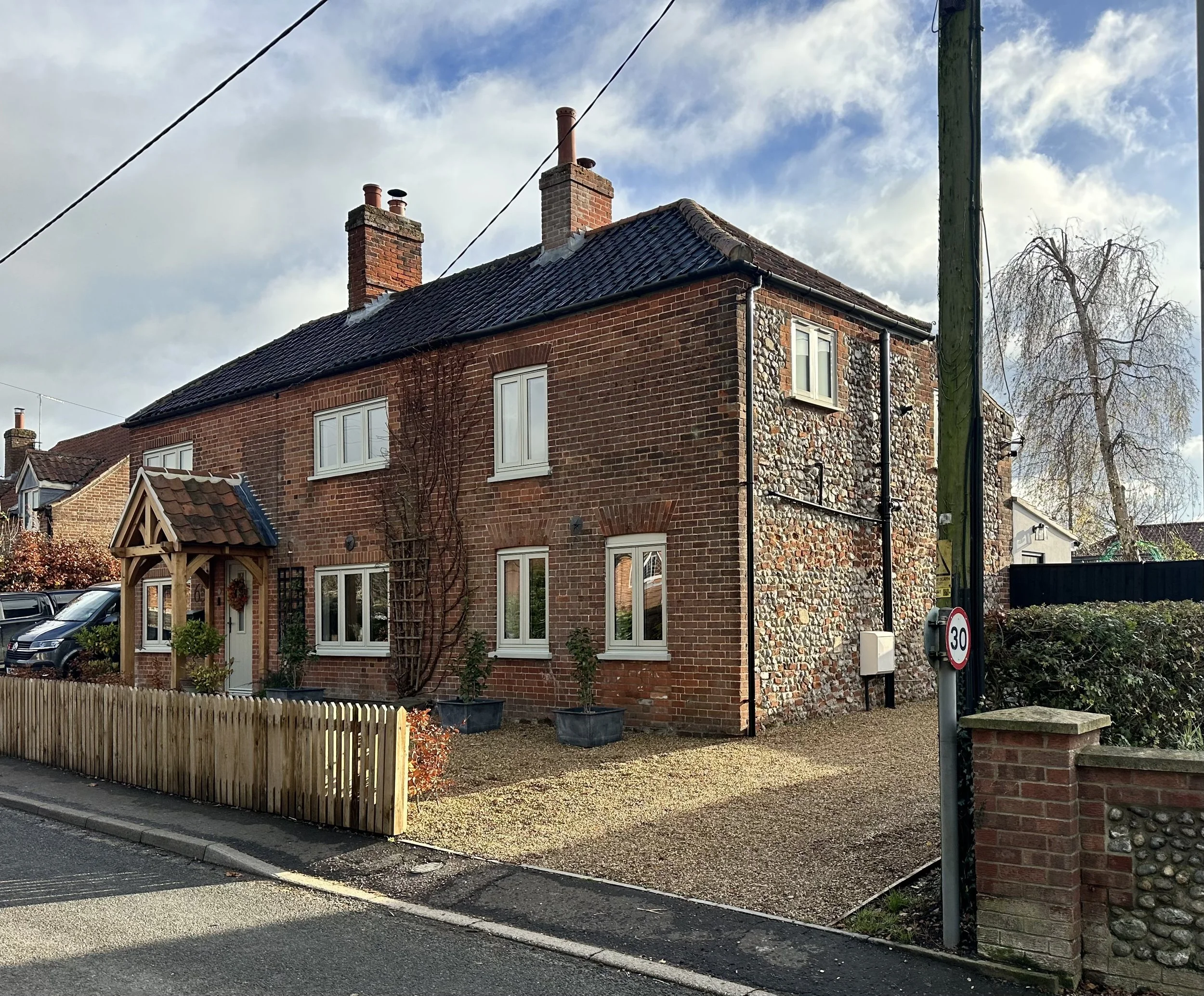 A brick house with a pitched roof, white window frames, and a small front yard with a wooden fence, potted plants, and a gravel pathway. There is a street sign indicating a speed limit of 30 mph and a utility pole with overhead wires. The sky is partly cloudy.