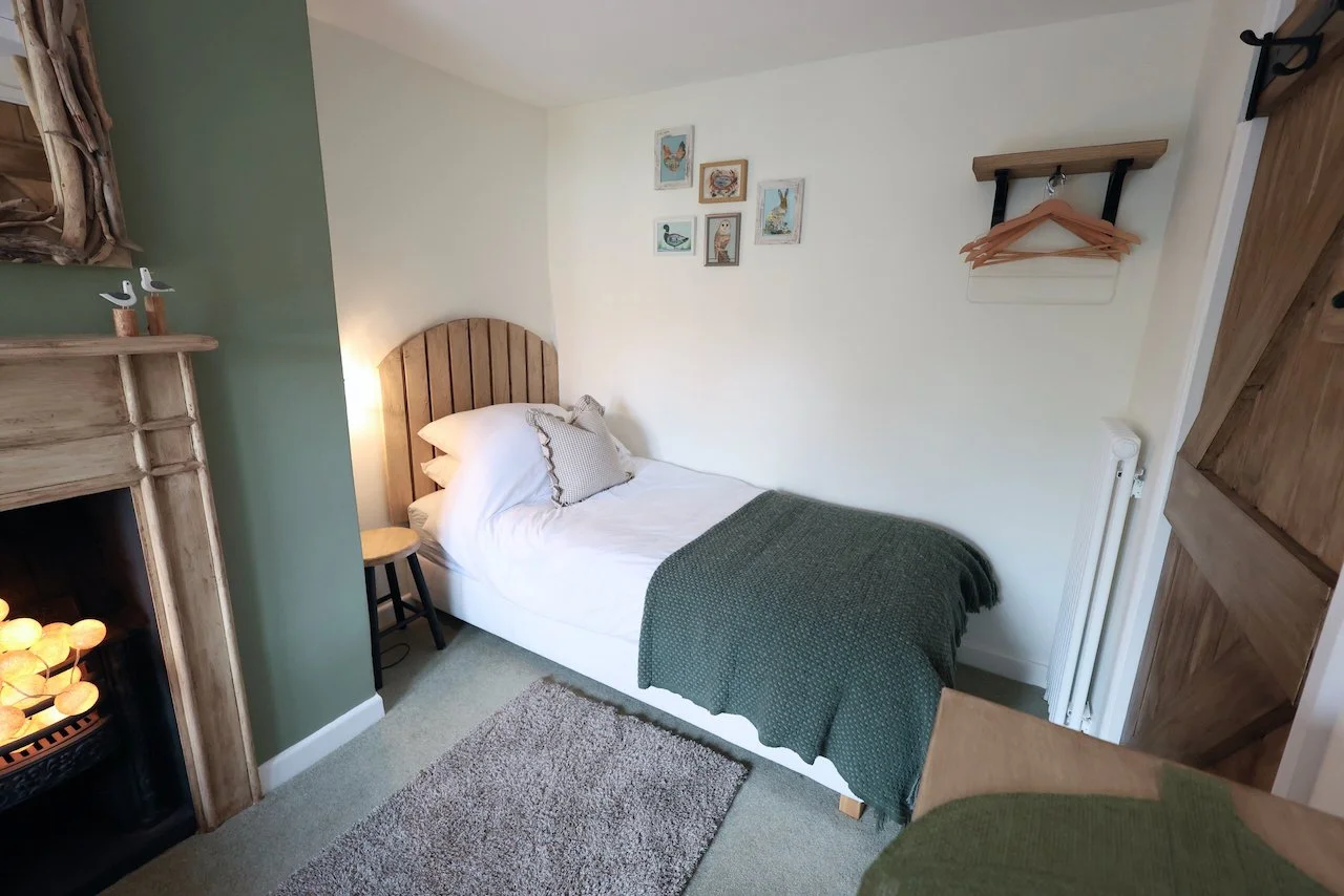 A cozy bedroom with a single bed, wooden headboard, white bedding, and a green throw blanket. There is a small chair beside the bed and framed bird artwork on the white wall. A rustic wooden shelf with hangers is mounted on the wall. A partial view of a fireplace with decorative lights and a green accent wall are visible boundary elements.