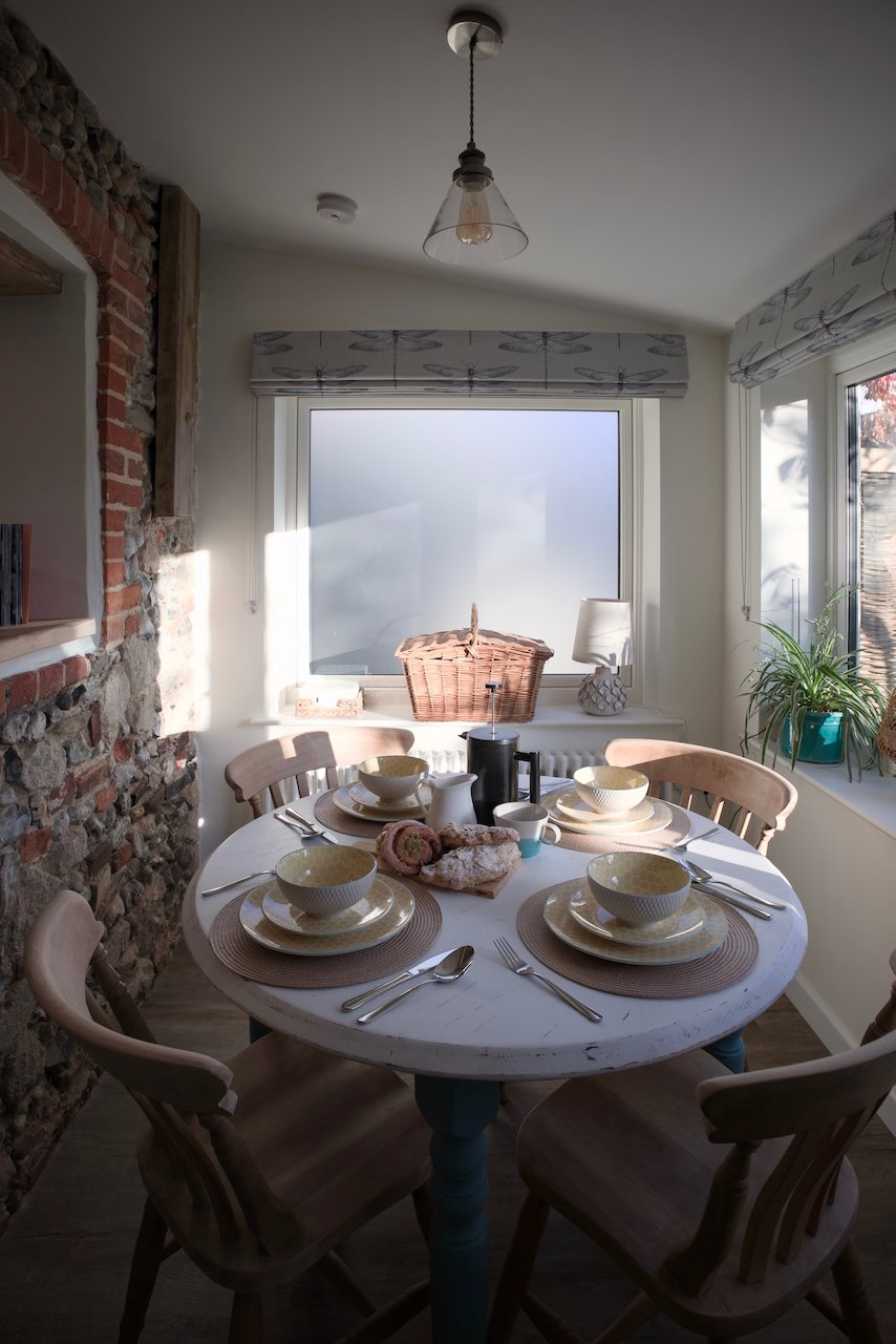 A cozy dining room with a round white table set with bowls, plates, cups, and utensils for five people. The table has a basket of bread, a teapot, and a small pitcher. Sunlight streams through frosted windows, with a brick wall on the left and housep