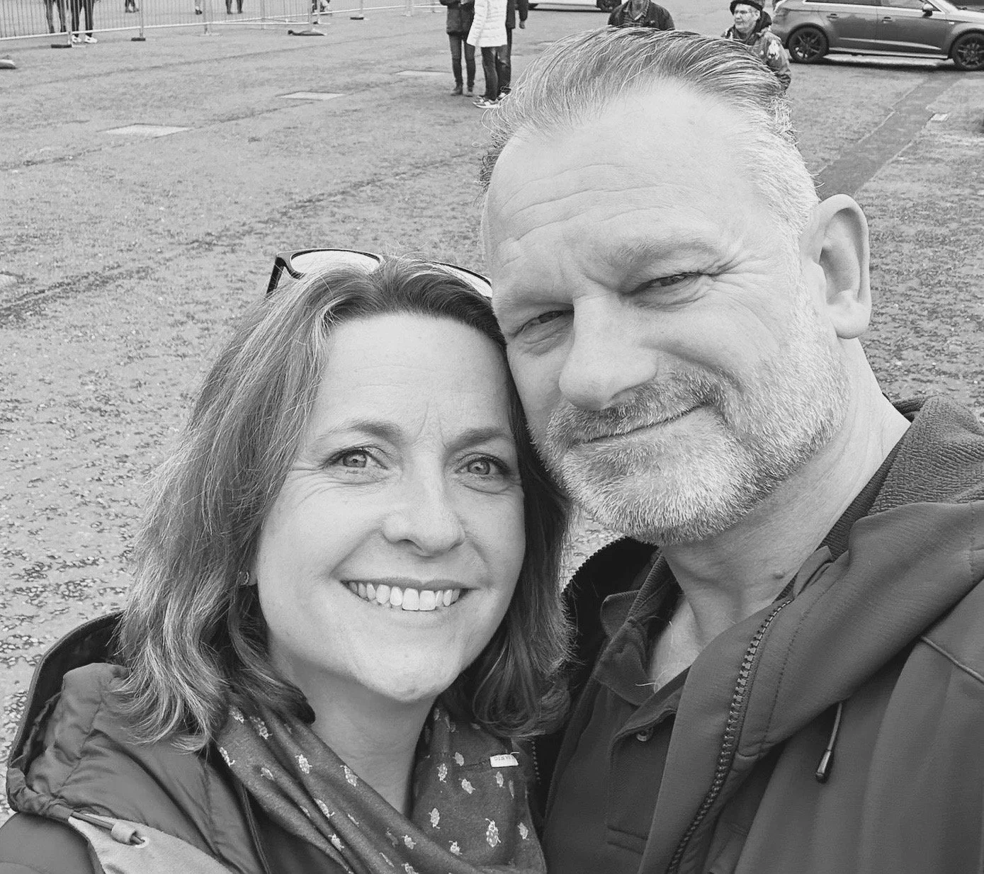 A black and white photo of a smiling woman and a man taking a selfie outdoors with a parking lot and a few people in the background.