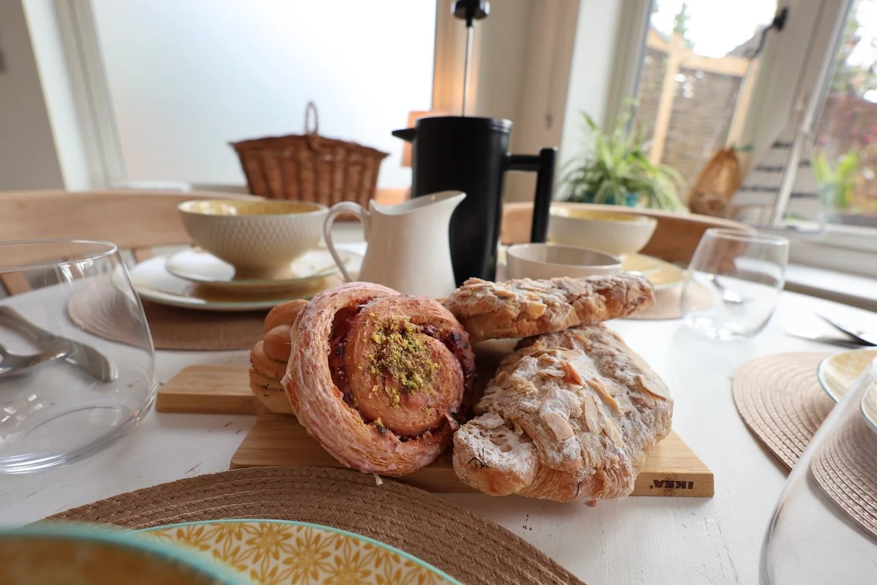 A breakfast table with pastries, bowls, and glasses, set in a bright dining room with plants and windows.