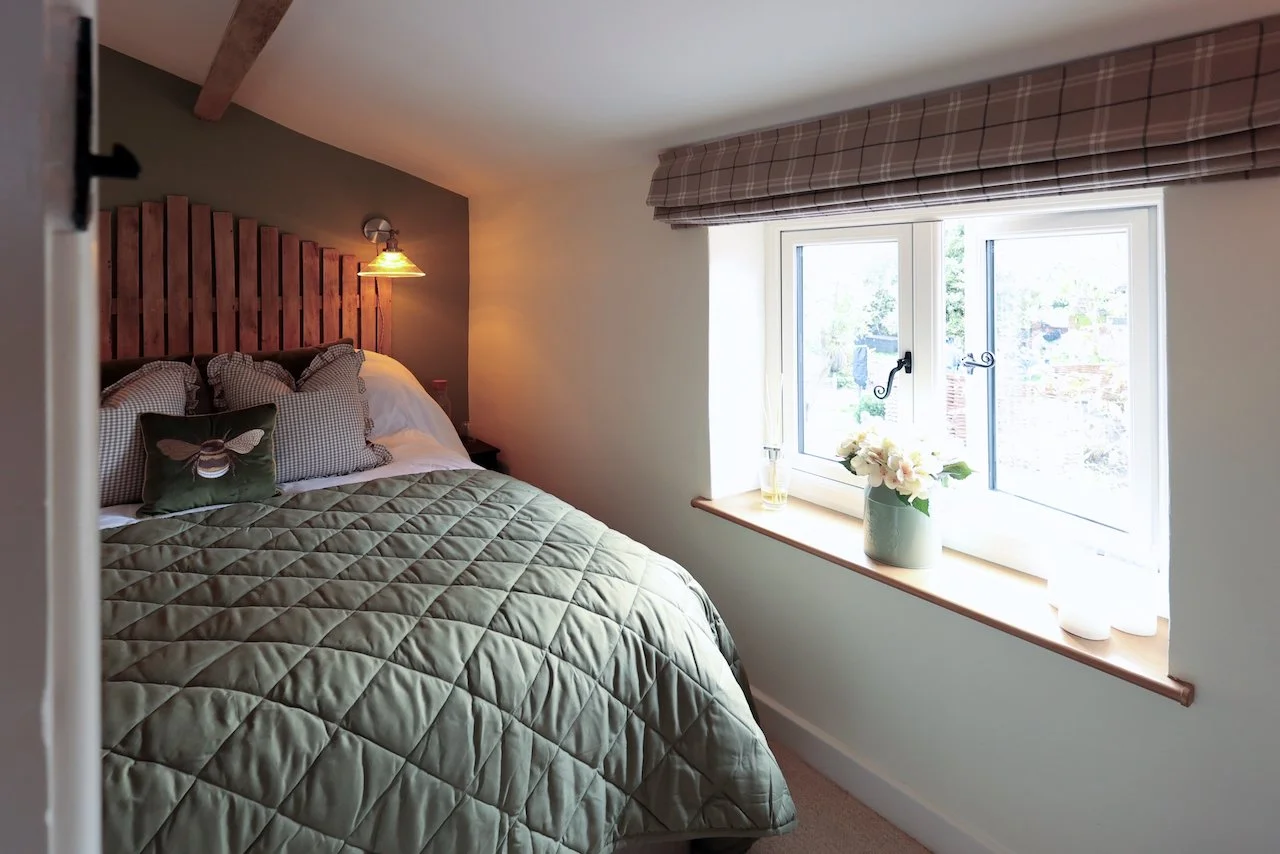 Comfortable bedroom with a bed, a wooden headboard, patterned pillows, a quilted green comforter, and a small wall-mounted lamp. There's a window with a plaid valance, a wooden windowsill with a vase of flowers, and a glass of water.