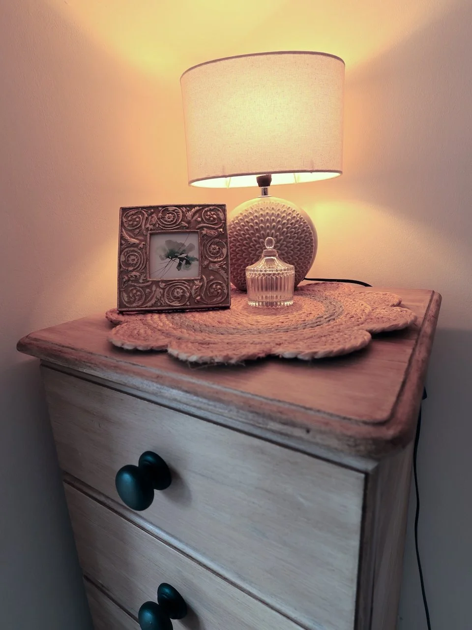 A wooden nightstand with two drawers, topped with a decorative doily, a framed photo, a textured table lamp with a beige shade, and a small glass container with a lid, all softly lit by the lamp.