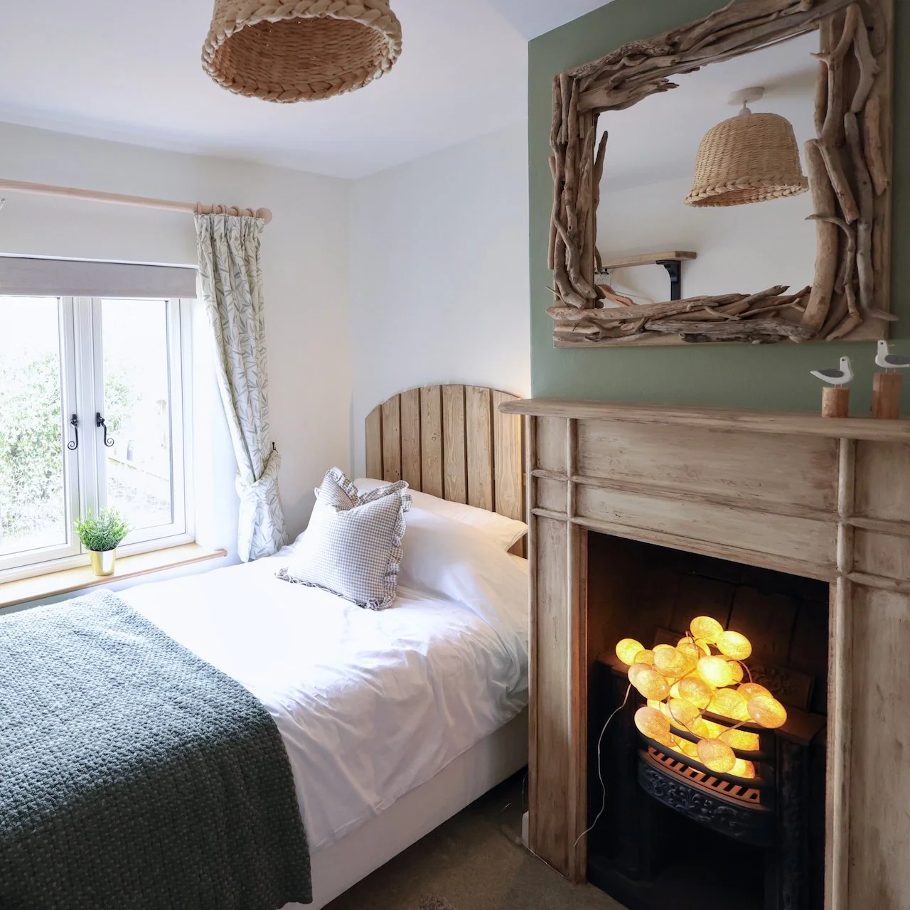 Cozy bedroom with a bed, striped pillows, a window with floral curtains, a plant, a driftwood mirror, a rustic fireplace with glowing decorative lights, and a wicker ceiling lamp.
