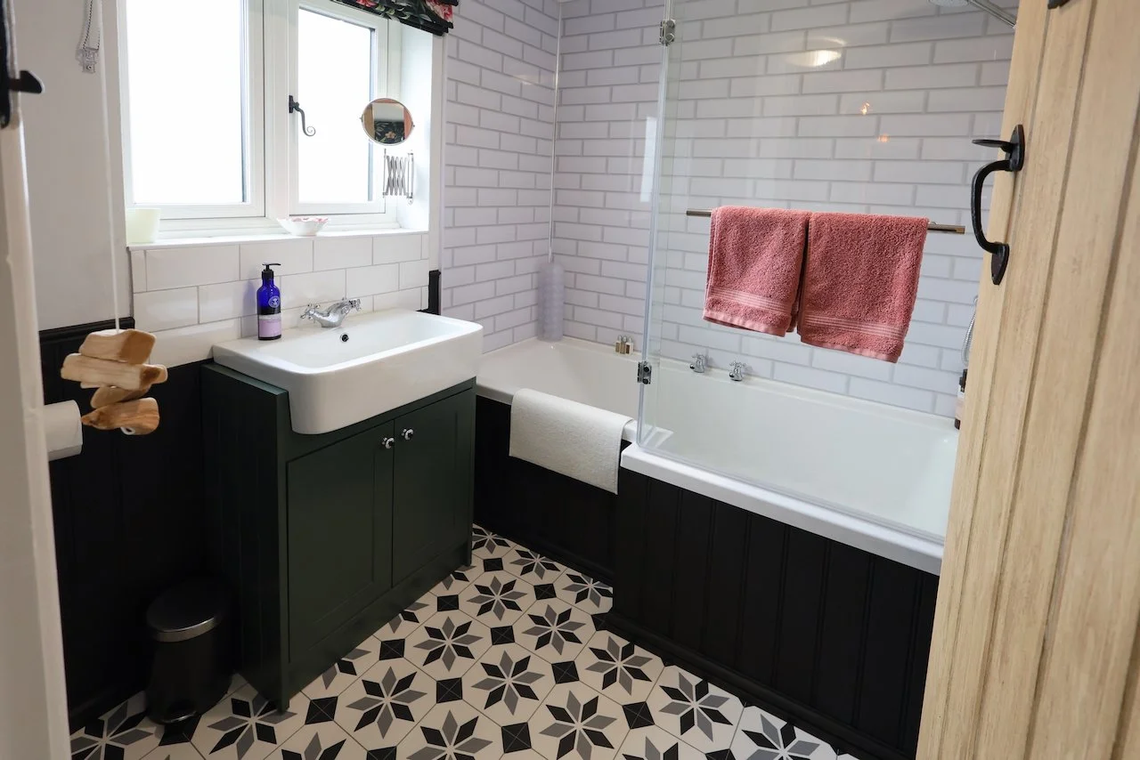 A bathroom with a white brick wall behind a bathtub, a dark green vanity with a white sink, patterned black and white tile floor, pink towels on a metal rod, a small mirror, a window, and a glass shower enclosure.