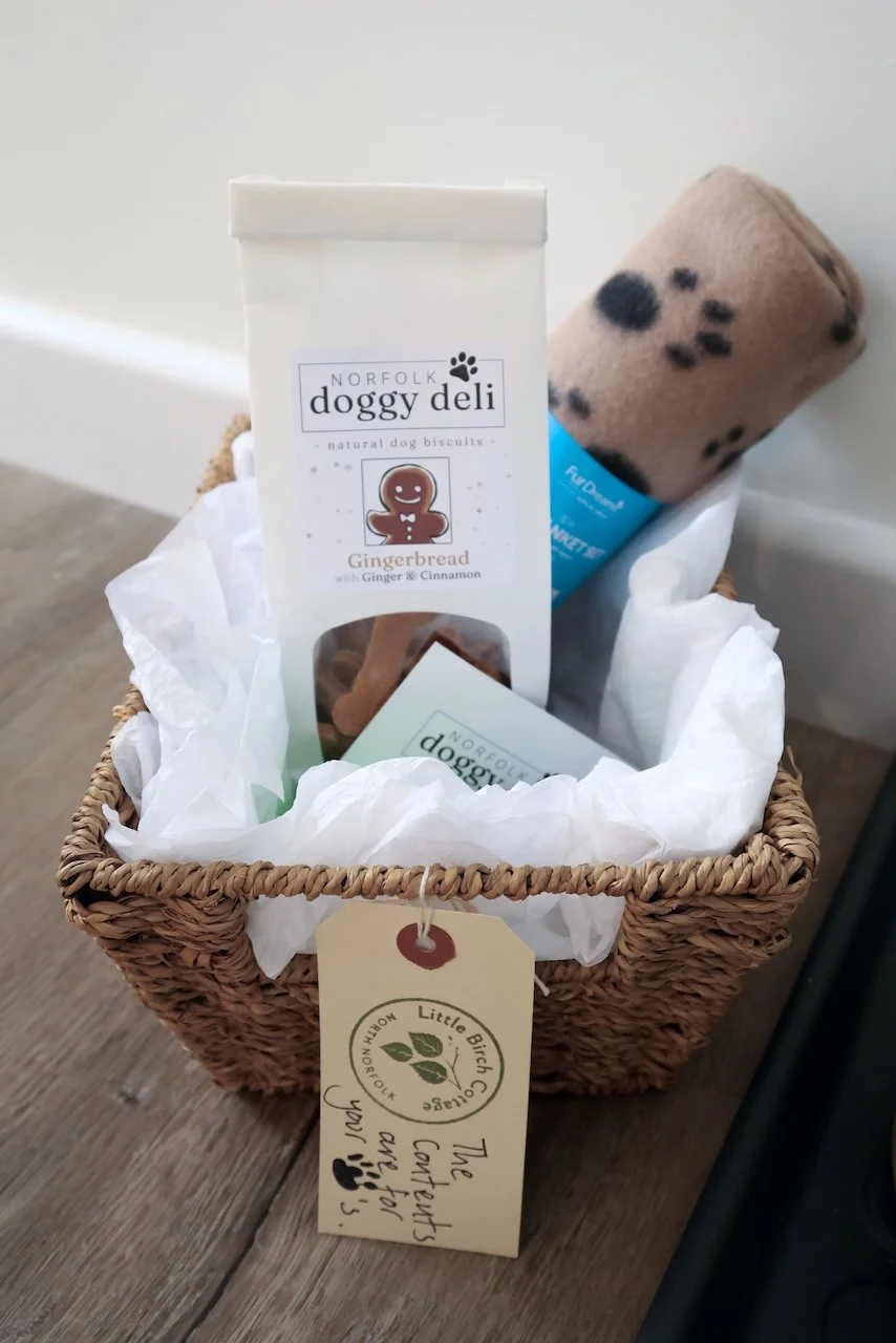 A wicker basket contains dog treats and a toy; the treats are Norfolk Doggy Deli gingerbread biscuits with ginger and cinnamon, and the toy appears to be a plush animal. A tag attached to the basket says 'The treats and toys are for your pup!'