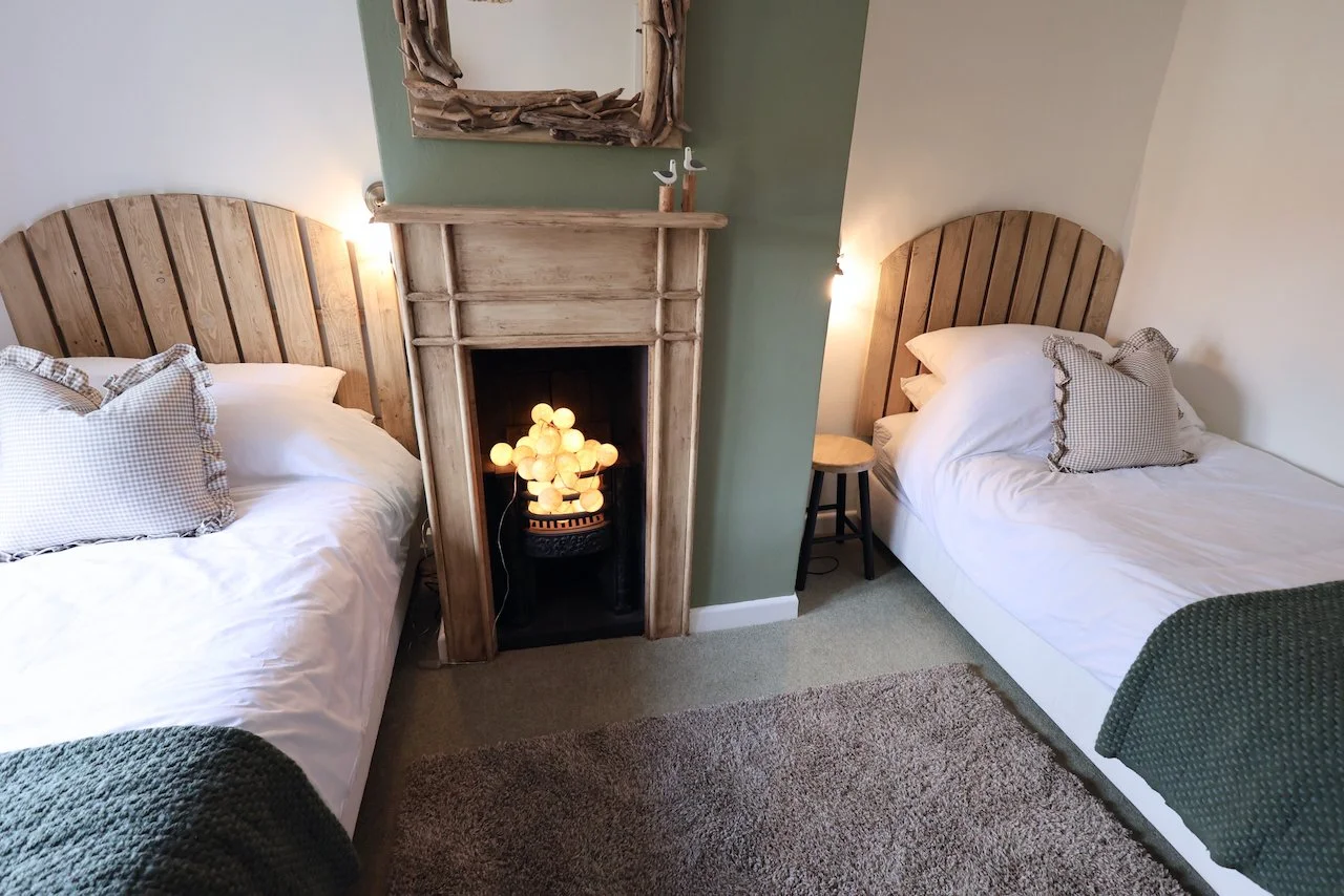 A cozy bedroom with two beds featuring wooden headboards, a fireplace with glowing string lights, a mirror above the fireplace, and a small stool beside one bed.