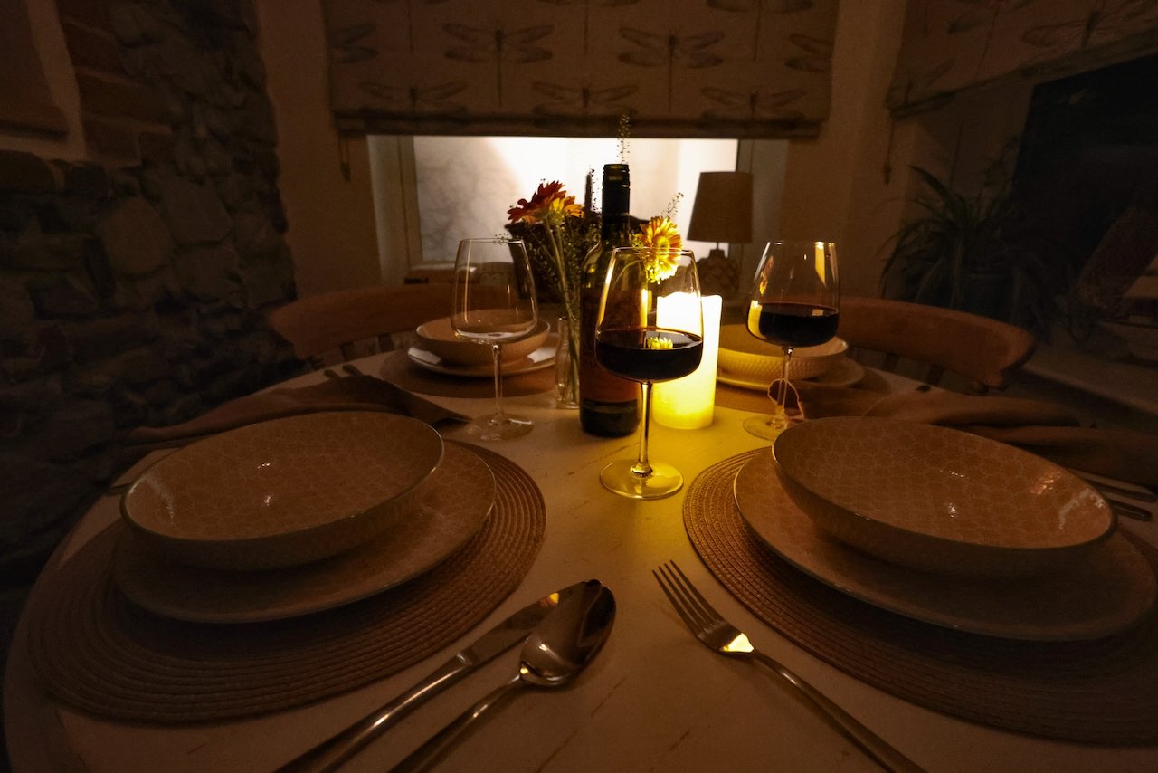 A dinner table set for four with plates, bowls, silverware, and glasses, illuminated by candles and a bottle of wine, in a cozy, dimly lit room.