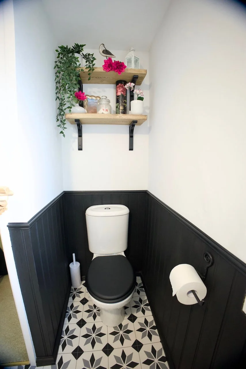 Small bathroom with black and white patterned tile floor, black wainscoting, white walls, a white toilet with black seat, toilet paper holder with roll of toilet paper, white toilet brush, and two wooden shelves above decorated with plants, flowers, a decorative bird figure, a lantern, and jars.