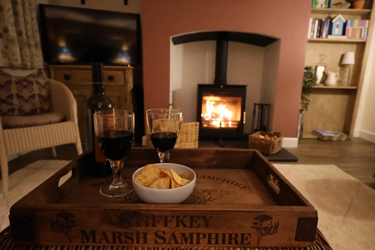 Living room with fireplace, two glasses of red wine, a bowl of potato chips on a wooden tray, a bottle of wine, and a television in the background.