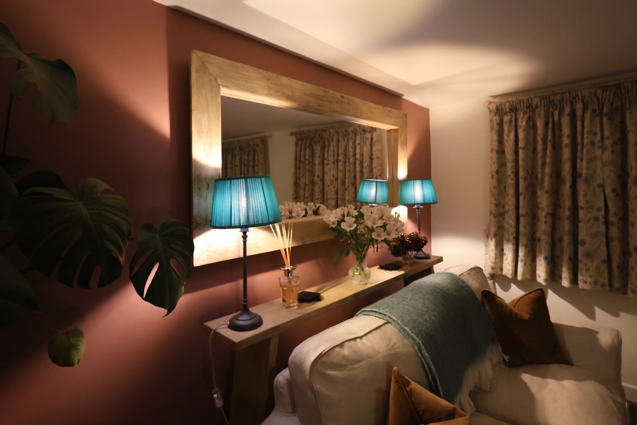 Living room with a large mirror, two teal lamps on a wooden console, a vase of flowers, a beige sofa with brown pillows, and floral curtains.