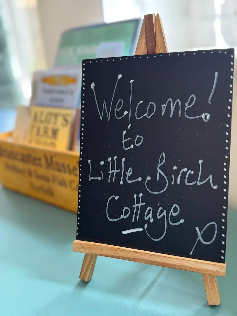 Small black chalkboard sign on a wooden easel with white handwritten text saying 'Welcome! to Little Finch Cottage'. In the background, blurred shelves with magazines or brochures on a teal surface.