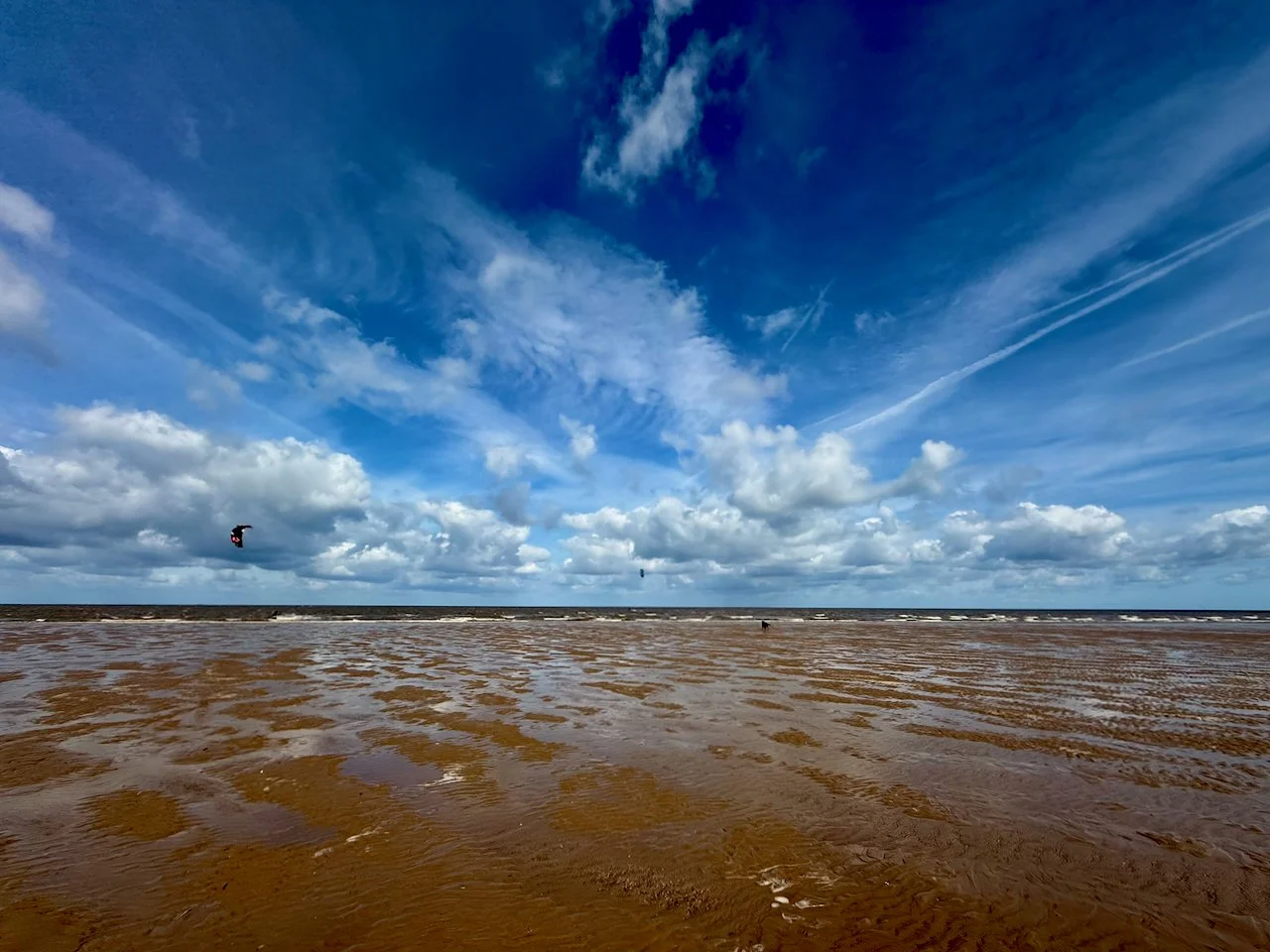 A wide view of a beach with wet sand, small waves, and a partly cloudy sky with contrails. Two kite surfers are seen in the distance, flying their kites.
