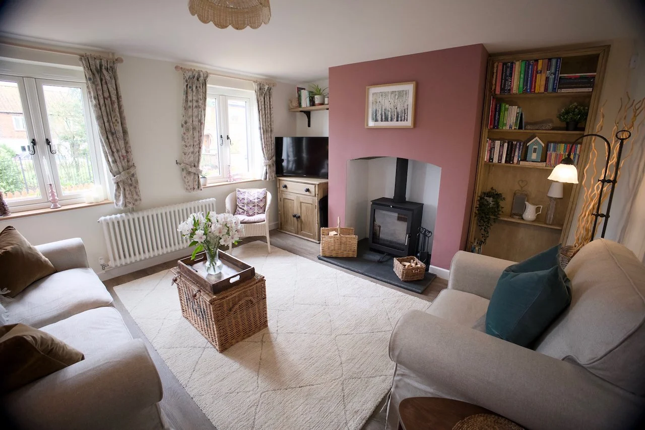 Living room with beige sofas, a wicker coffee table with flowers, a fireplace with a pink wall, a bookshelf, and a window with floral curtains.