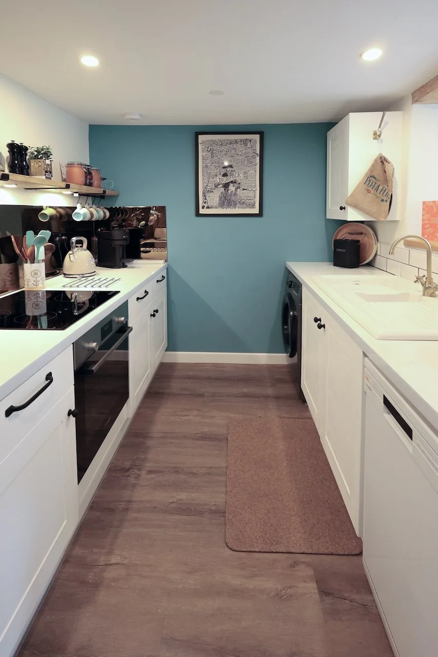 A compact kitchen with white cabinets, a brown rug, and wood flooring. The left side has a stovetop, kettle, and utensils, while the right side features a sink, a washing machine, and a bread box. A blue accent wall has a framed artwork, and there are jars and decorations on shelves.
