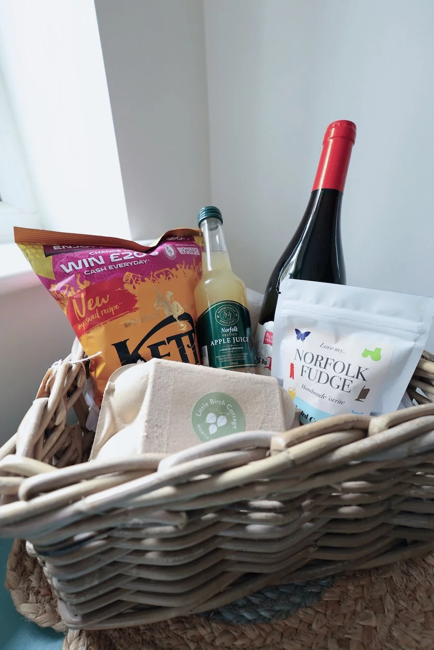 A wicker basket filled with various items including a packet of Kez brand food, a bottle of Norfolk apple juice, a bottle of red wine, a bag of Norfolk fudge, and a container labeled Little Birch Cottage.