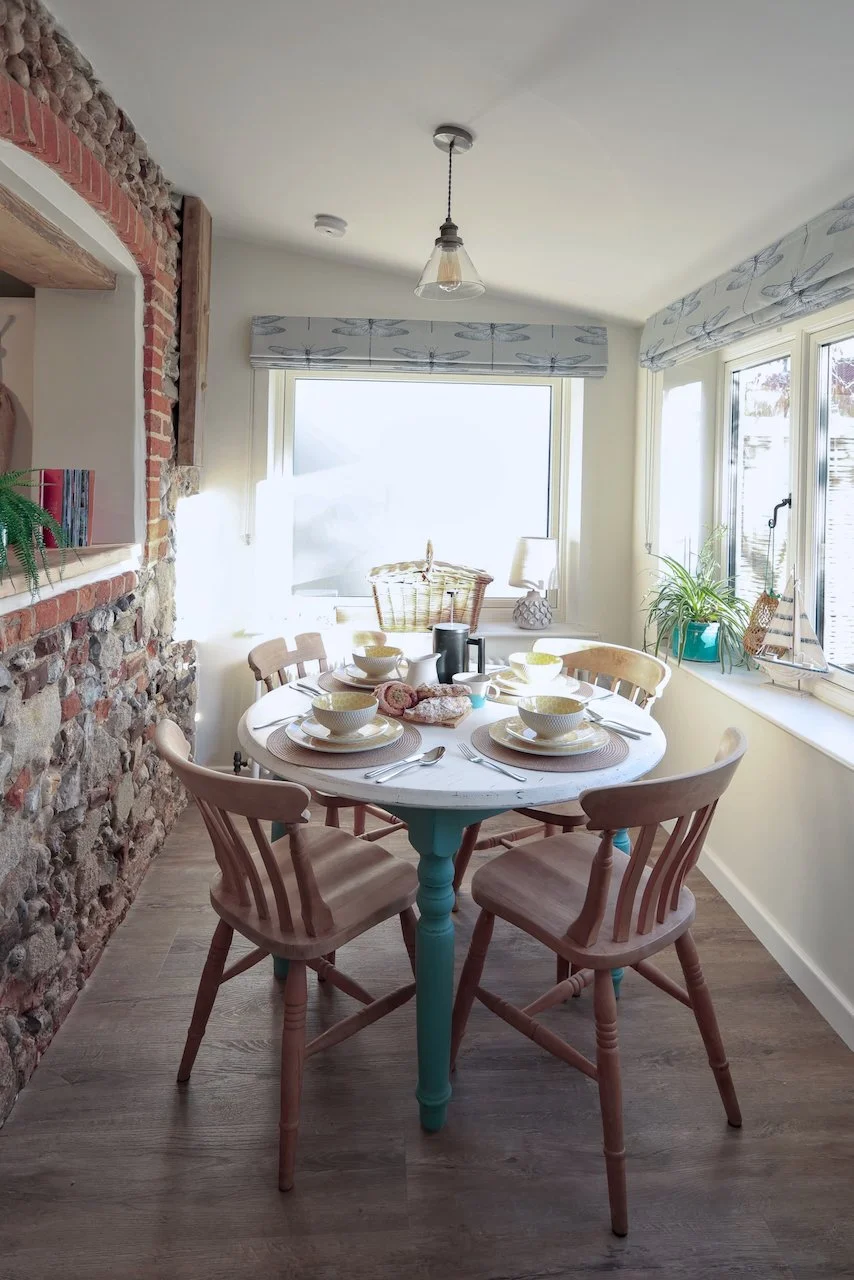 A dining area with a round white table set for six, surrounded by wooden chairs with beige cushions. The table has bowls, plates, utensils, and napkins. A large window behind the table lets in natural light. There is a brick wall on the left and a windowsill with plants and decorative items on the right.