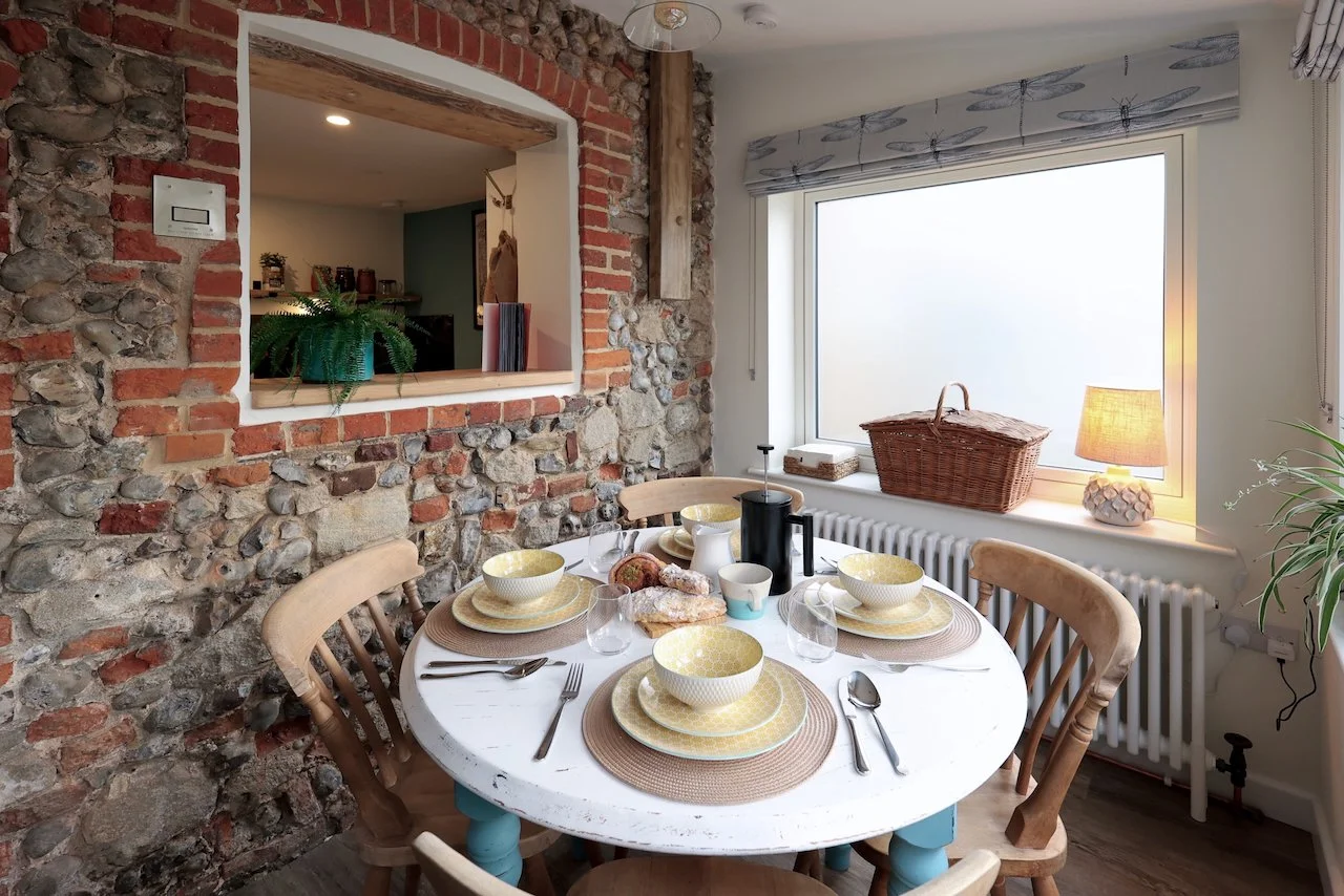 Dining room with a round white table set with yellow and brown bowls and plates, silverware, and glasses. Four wooden chairs surround the table. A window with a dragonfly-patterned blind is in the background, with a wicker basket and a small lamp on the windowsill. A brick and stone wall with a window opening is on the left side of the image.