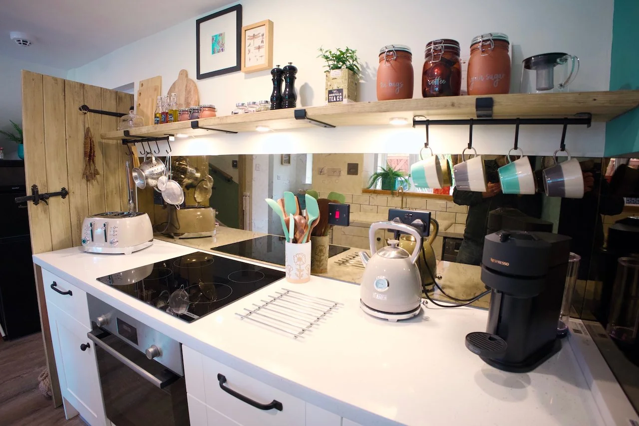 Modern kitchen with white countertop, electric cooktop, white oven, and various kitchen appliances including a toaster, kettle, and coffee machine. Open wooden shelves hold jars, cups, and decorative items. Hanging hooks hold utensils and mugs. There's a mirror on the wall reflecting part of the room.