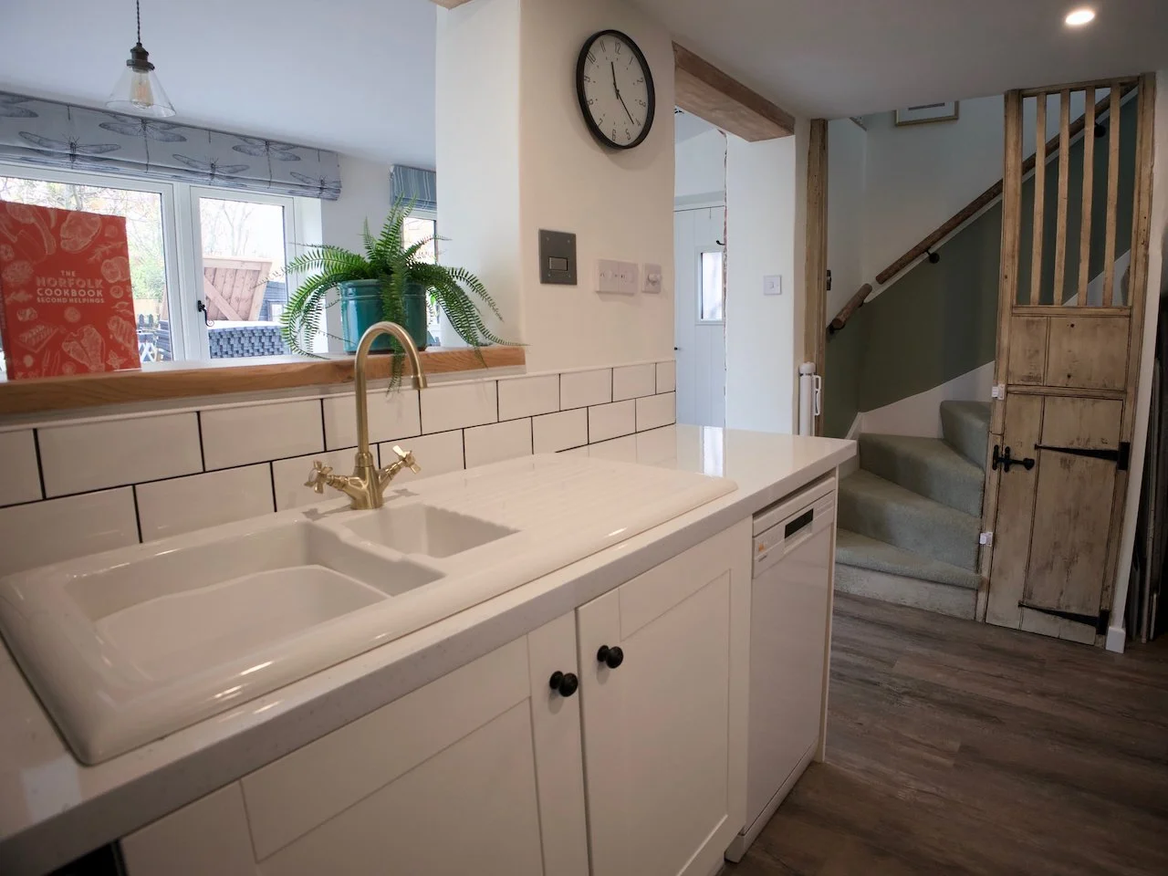 Kitchen with white sink, brass faucet, white cabinets, wood countertop, green plant in teal pot, window with blinds, staircase with beige carpet, wooden door, and a wall clock showing 4:55.